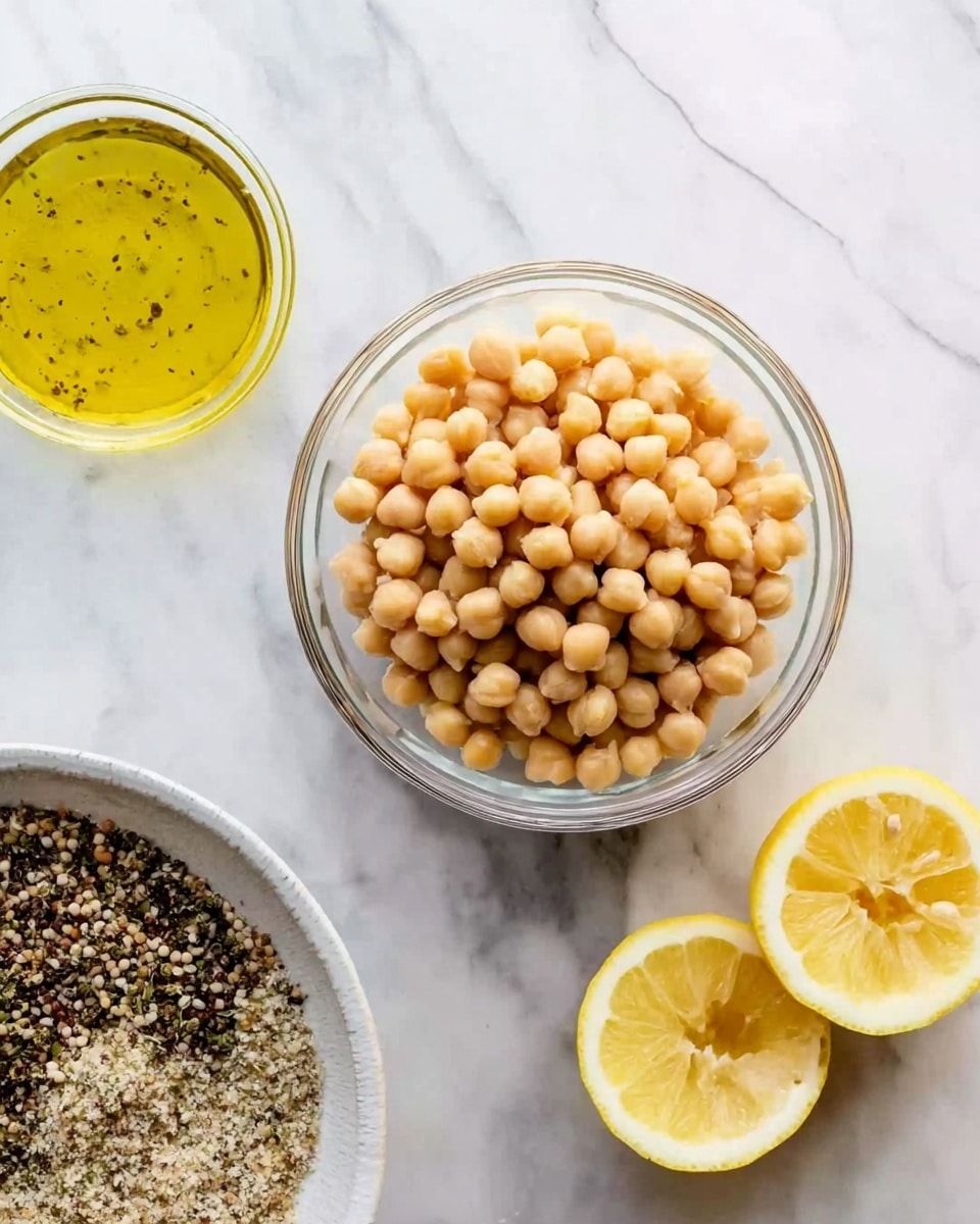 A clear glass bowl filled with pale beige chickpeas sits near the center on a white marbled surface. To the top-left of the bowl, there is a small glass container filled with a yellow liquid that looks like olive oil with seasonings. On the right side, two lemon halves with a bright yellow color rest directly on the white marbled surface. At the bottom of the image, part of a white bowl is visible, filled with a coarse mixture of black, white, and brown grains or seeds. The whole scene is bright with soft natural light, showing clear, simple textures and colors. photo taken with an iphone --ar 4:5 --v 7