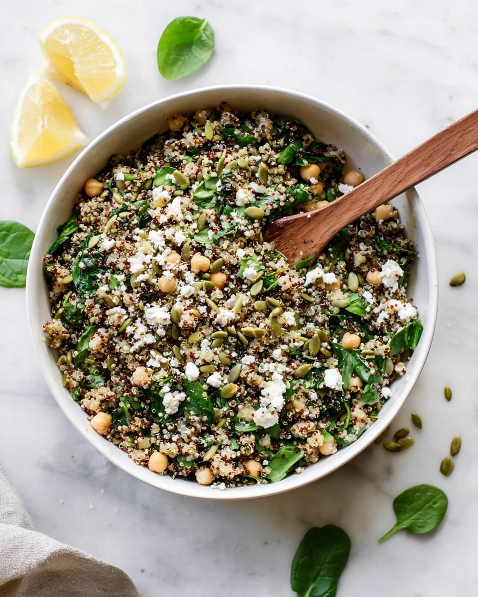 A white bowl sits on a white marbled surface filled with a mixed salad of three visible layers: a base of cooked quinoa with small grains in white, red, and black colors, mixed throughout with bright green spinach leaves that add a fresh texture, and topped with scattered light beige chickpeas and pale green pumpkin seeds. Crumbled white cheese is sprinkled over the top, adding a soft, crumbly texture. A wooden spoon stands upright in the bowl, ready to stir the mix. To the left, two squeezed lemon halves and a spinach leaf rest on the white marbled surface. photo taken with an iphone --ar 4:5 --v 7
