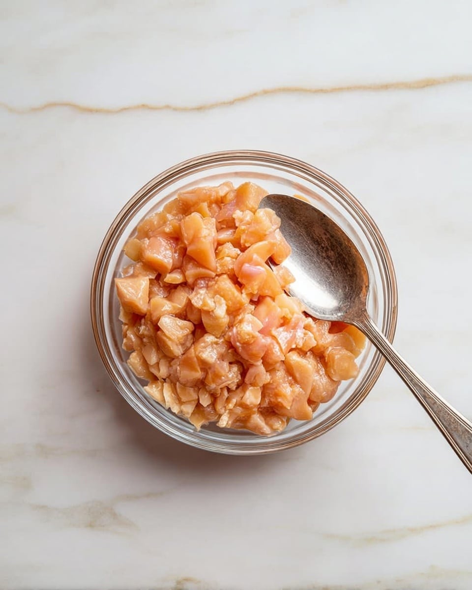 A clear glass bowl filled with small, shiny pieces of raw chicken, light pink and orange in color, with a silver spoon resting inside on the right side of the bowl. The bowl is placed on a white marbled surface. photo taken with an iphone --ar 4:5 --v 7