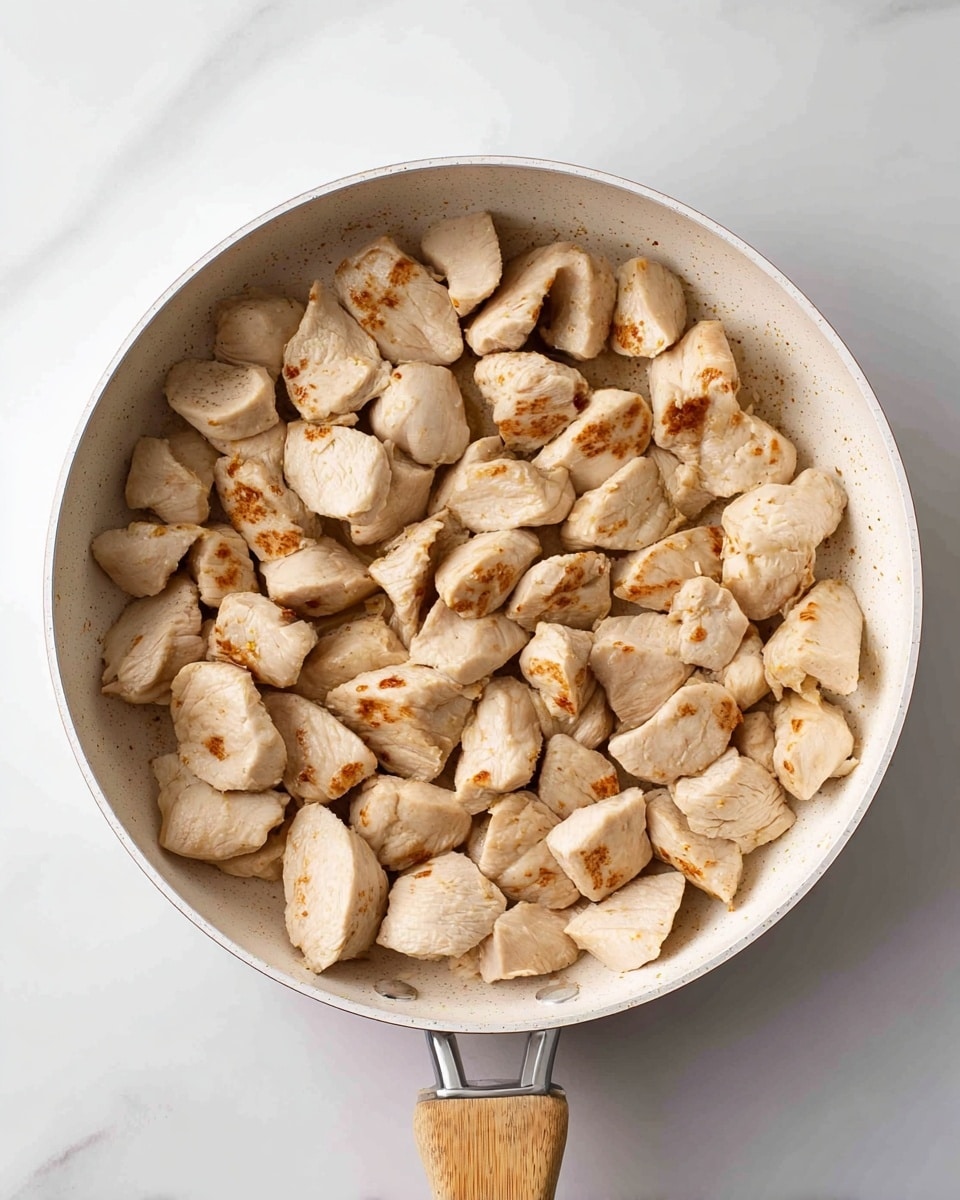 A white pan with a light wooden handle is filled with pieces of cooked chicken. The chicken pieces are light beige with slight brown grill marks on the surface, showing they are cooked but not browned fully. The pieces vary in size and are unevenly shaped, filling the pan almost to the top. The pan sits on a white marbled surface, creating a clean and bright background. photo taken with an iphone --ar 4:5 --v 7