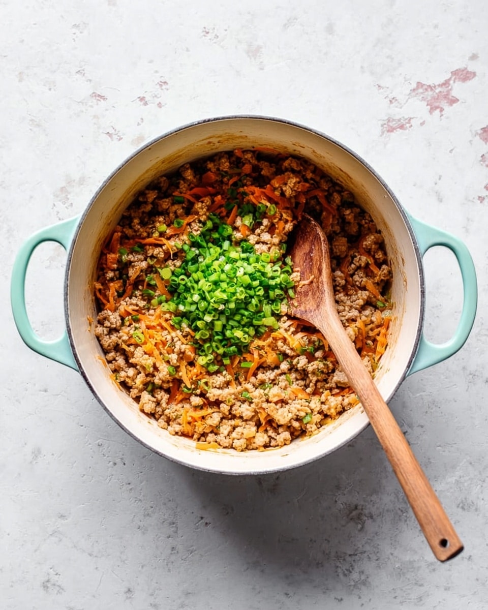 A top view of a turquoise handled white pot filled with a cooked mixture of light brown crumbled meat and orange carrot shreds mixed evenly inside. On top of this mixture, there is a small mound of bright green chopped scallions placed right in the center. A wooden spoon with a long handle rests inside the pot, its handle extending outward to the bottom right edge. The pot is on a white marbled surface with subtle grey and pink marks. photo taken with an iphone --ar 4:5 --v 7