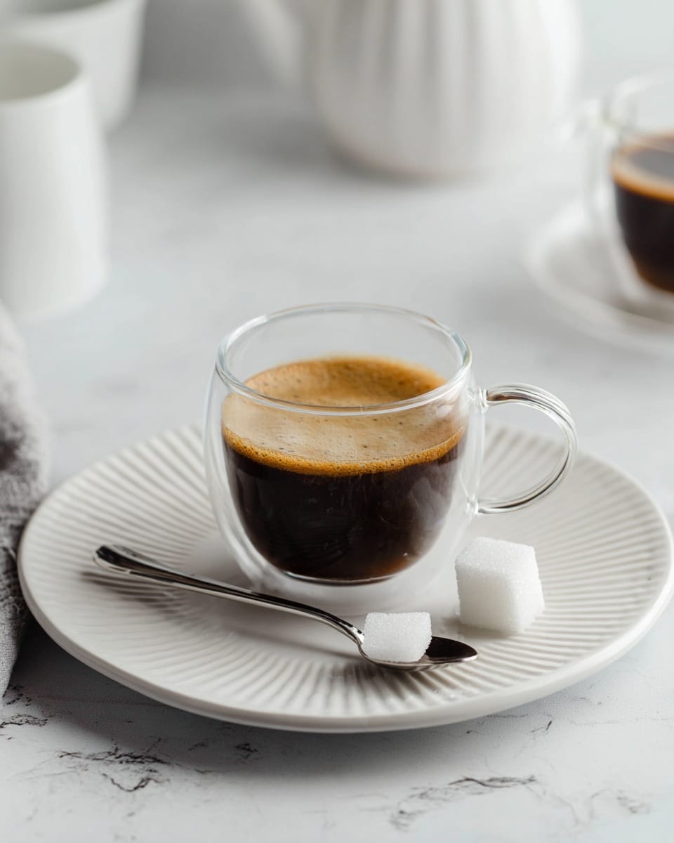 A clear glass cup with a handle is filled halfway with dark brown coffee that has a light foam layer on top. The cup sits on a large white plate with a ridged edge pattern. On the plate next to the cup, there is a shiny silver spoon holding a white sugar cube. The surface underneath is a white marbled texture, and in the background, there are blurred white ceramic items and another glass cup with coffee partially visible. The lighting is soft, giving a clean and calm look. photo taken with an iphone --ar 4:5 --v 7