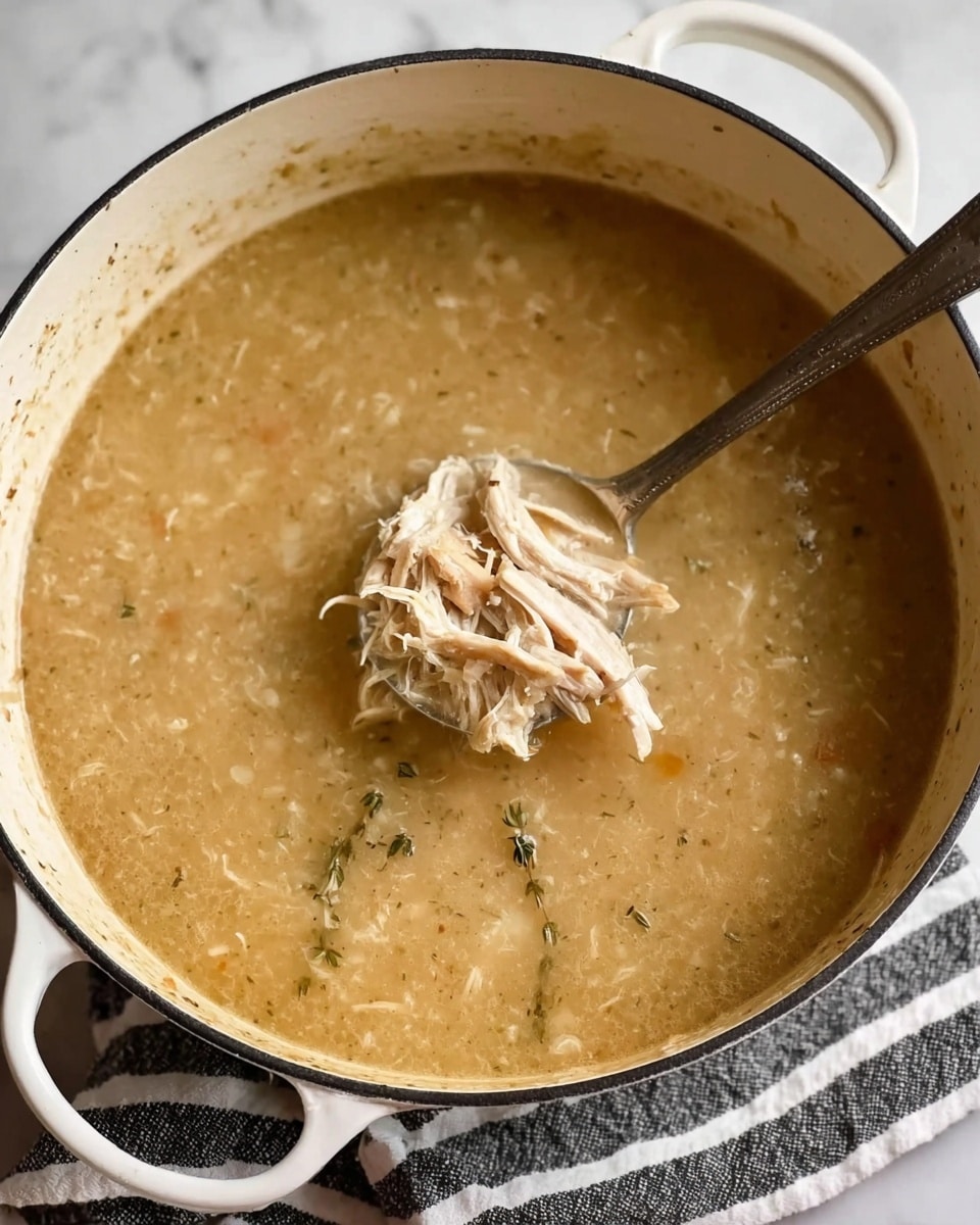 A white pot filled with thick soup that is light brown and slightly creamy in texture, with visible small bits of herbs and spices. In the center, there is a spoon scooping up shredded pale yellow meat from the soup. The pot rests on a white marbled surface with a black-and-white striped cloth partially visible underneath. The overall look is warm and comforting, showing a rich, homemade soup. photo taken with an iphone --ar 4:5 --v 7
