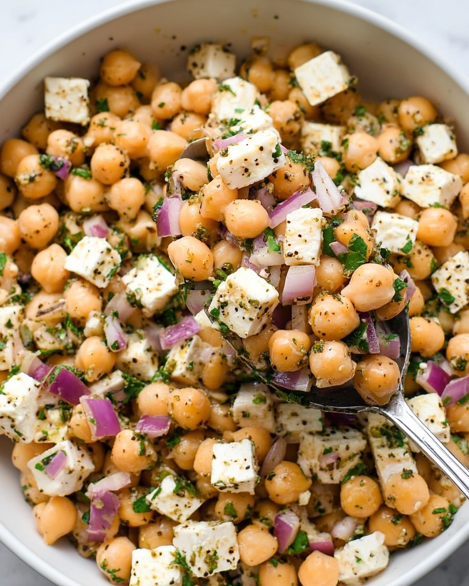 A close-up view of a white bowl filled with a chickpea salad that has three main layers: round, light orange chickpeas form the base, mixed throughout with small cubes of white feta cheese and finely chopped purple onion pieces, all sprinkled with dark green chopped parsley and black pepper, visible as tiny specks; in the center, a spoon holds a scoop of this mixture, showing the colorful textures closely against a white marbled surface. photo taken with an iphone --ar 4:5 --v 7