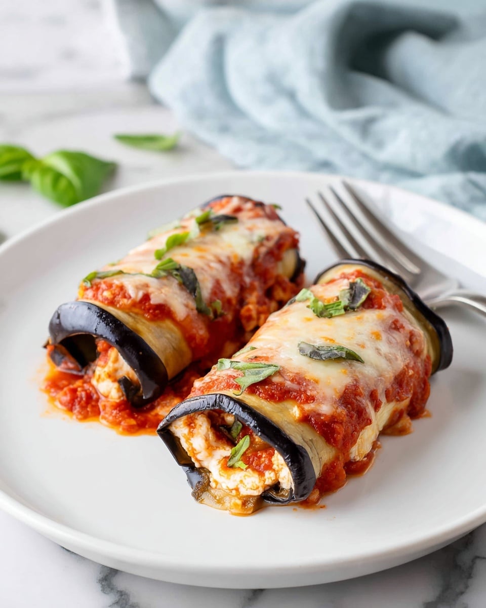 Two eggplant rolls sit side by side on a white plate with a silver fork behind them. Each roll has a deep purple eggplant outer layer, filled with a creamy, light beige mixture that is visible at the ends. The top of each roll is covered with bright red tomato sauce layered with melted, slightly golden cheese and garnished with small fresh basil leaves. The plate is placed on a white marbled surface with a light blue cloth slightly visible in the background. Photo taken with an iphone --ar 4:5 --v 7