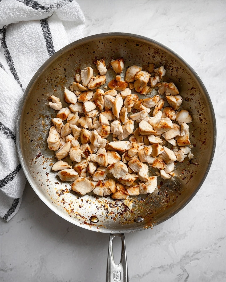 A metal pan filled with small pieces of cooked chicken, mostly white with light golden-brown edges scattered unevenly across the surface. The pan shows some dark browned spots from cooking, and it rests on a white marbled surface. To the top left of the pan, there is a white towel with black stripes casually placed. Photo taken with an iphone --ar 4:5 --v 7