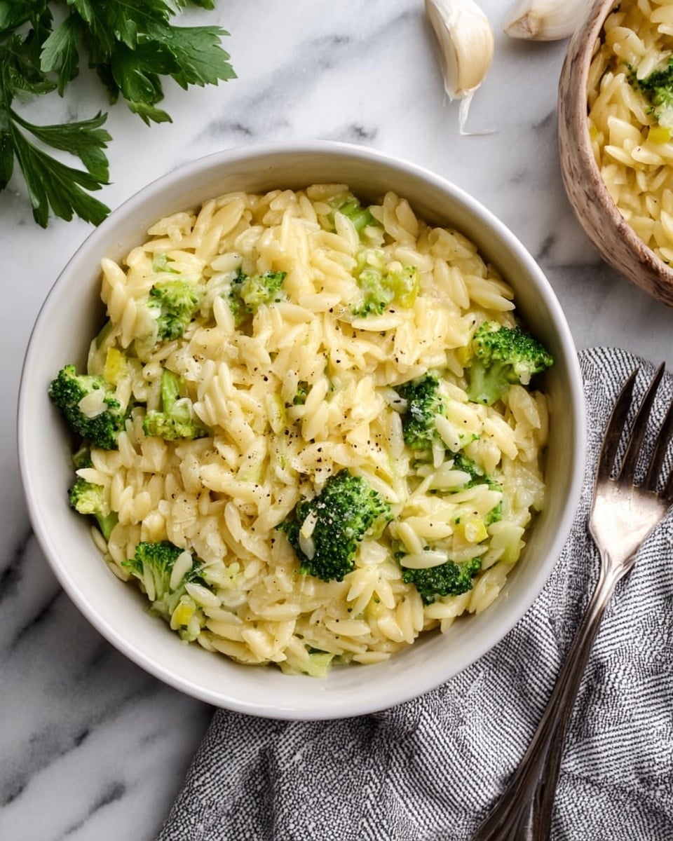 A white bowl filled with creamy yellow orzo pasta mixed with bright green broccoli florets, with small bits of melted cheese or onion spread throughout. The orzo looks soft and coated in a smooth sauce, and some black pepper is sprinkled on top. The bowl sits on a white marbled surface with a gray and white striped cloth napkin to the right, next to a silver fork. There are also green parsley leaves to the left and a bowl of garlic cloves toward the top right corner. photo taken with an iphone --ar 4:5 --v 7