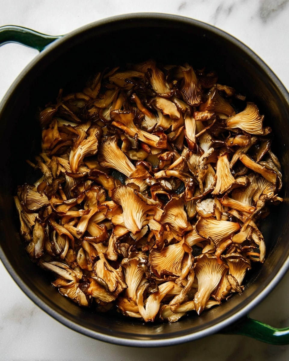 A top view of a large black pot filled with a single layer of cooked Maitake mushrooms, showing their soft, curled shapes and a mix of golden brown and dark brown colors from cooking. The mushrooms have a slightly shiny texture and cover the bottom of the pot evenly. The pot is set against a white marbled surface, with a green handle visible on one side. Photo taken with an iphone --ar 4:5 --v 7