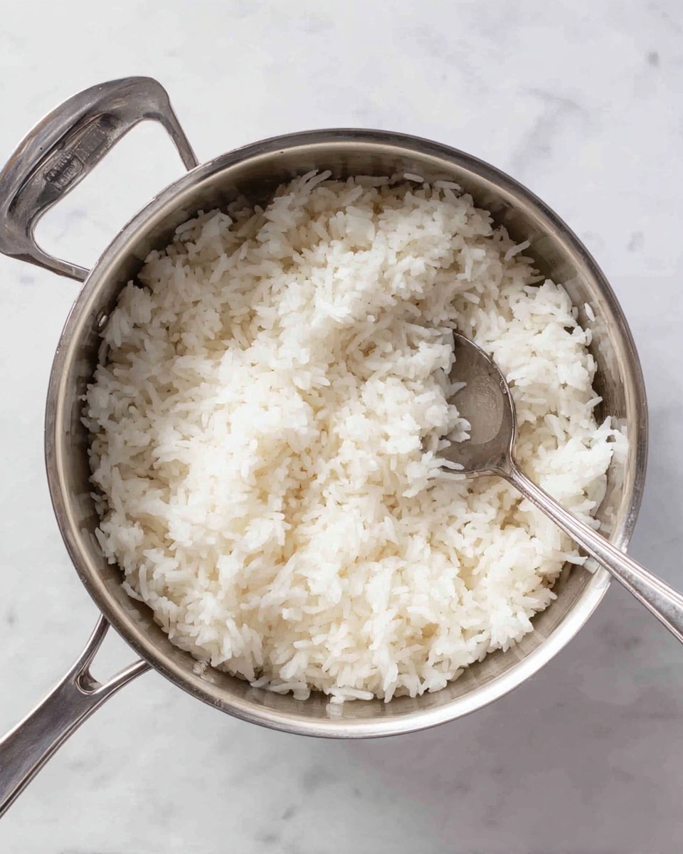 The image shows a silver metal pot filled with white cooked rice. The rice looks soft and fluffy, filling the pot evenly with visible clumps but no dryness. A silver spoon is placed inside the pot resting on the rice, with the handle pointing towards the right side. The pot is placed on a white marbled surface, and the pot lid with a silver handle lies upside down nearby to the left. Photo taken with an iphone --ar 4:5 --v 7