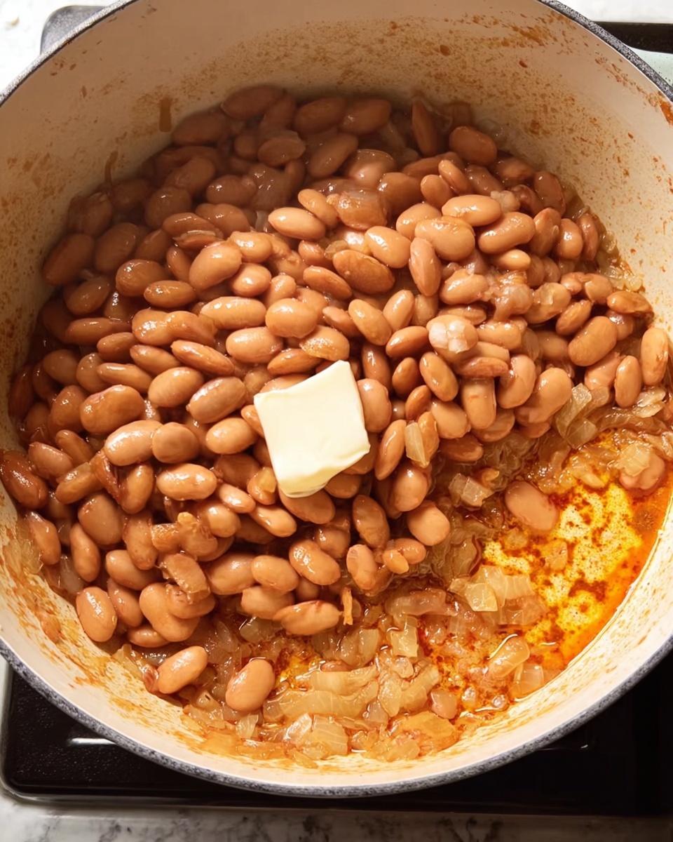 A white cooking pot filled with layers of cooked light brown beans on top, with a small square of white butter slightly melted in the middle. At the bottom layer, golden brown sautéed onions and garlic mix with a reddish-orange oily sauce that covers the surface of the pot. The pot sits on a stove, with a hint of black around the edges. The white marbled surface is visible around the pot's base. photo taken with an iphone --ar 4:5 --v 7