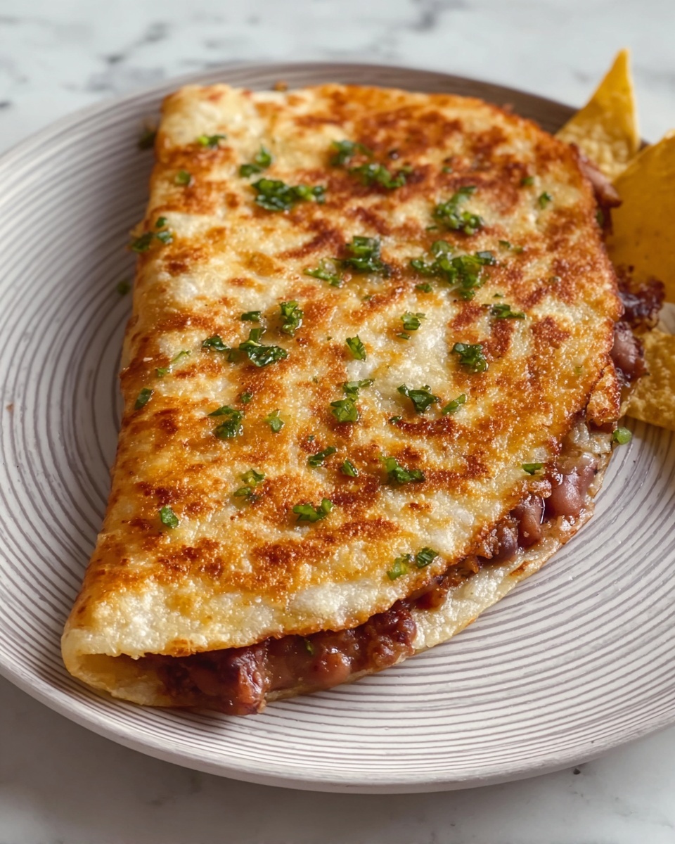 A single folded white tortilla with a golden-brown crispy surface on top sprinkled with small pieces of chopped green herbs. Inside the tortilla, a layer of soft, reddish-brown refried beans is visible around the edges. The tortilla rests on a round white plate with a subtle gray striped pattern, placed on a white marbled surface. A small part of a yellow tortilla chip is visible near the plate. Photo taken with an iphone --ar 4:5 --v 7