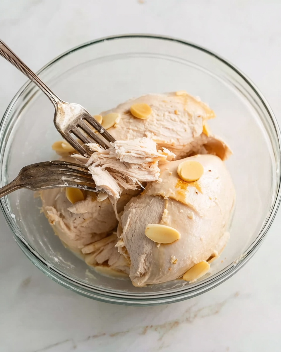 A clear glass bowl holds three large pieces of light beige cooked chicken with some visible circular slices of cooked garlic on top. Two forks are placed inside the bowl, one held by a woman's hand reaching in from the left side, shredding one piece of chicken, revealing a slightly fibrous texture inside. The bowl is set on a white marbled surface with soft natural lighting that highlights the textures and colors clearly. photo taken with an iphone --ar 4:5 --v 7
