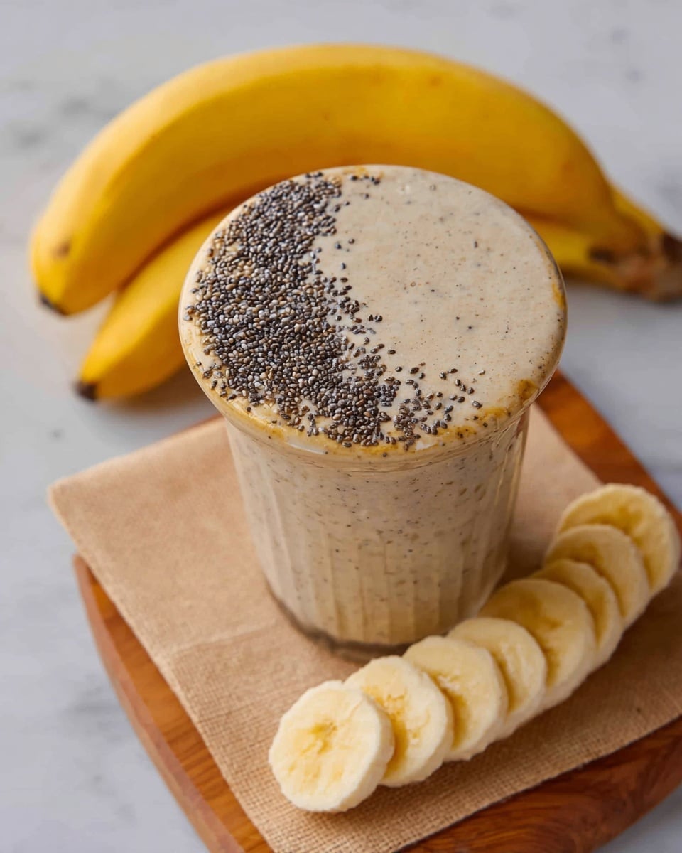 A clear glass filled with a thick, light beige smoothie topped with small black chia seeds mostly on one side of the surface, with some smoothie spilled slightly over the rim showing its creamy texture. The glass sits on a wooden board with a beige napkin underneath, next to a row of seven evenly spaced pale yellow banana slices with soft smooth texture. In the background, two whole yellow bananas with dark spots rest on a white marbled surface photo taken with an iphone --ar 4:5 --v 7