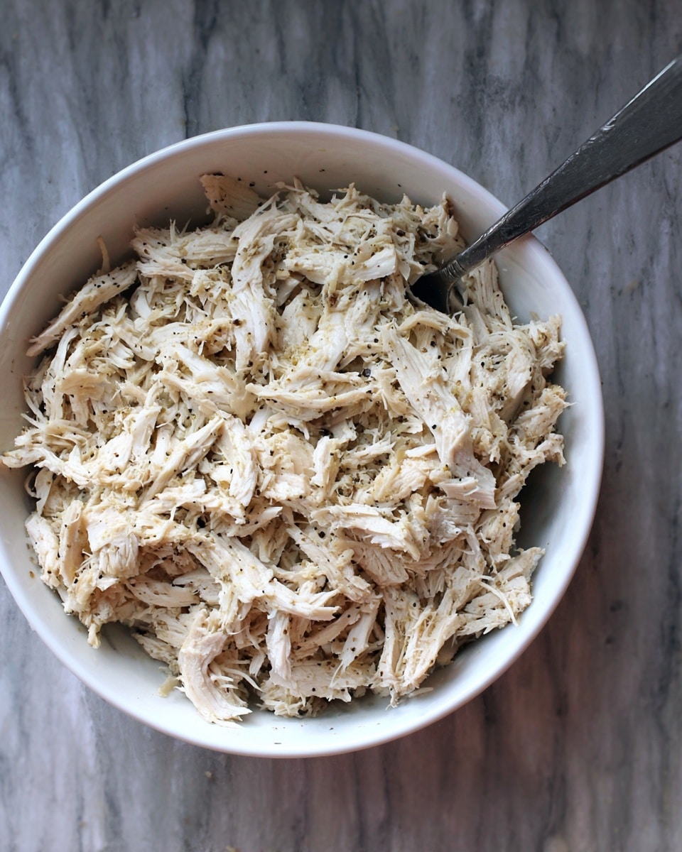 The image shows a round white bowl filled with shredded white meat mixed with small black specks, suggesting seasoning. The shredded pieces are soft and fibrous with a moist texture, piled loosely in the bowl. A silver spoon is placed inside, partially buried in the meat. The bowl sits on a surface with a white marbled texture. photo taken with an iphone --ar 4:5 --v 7