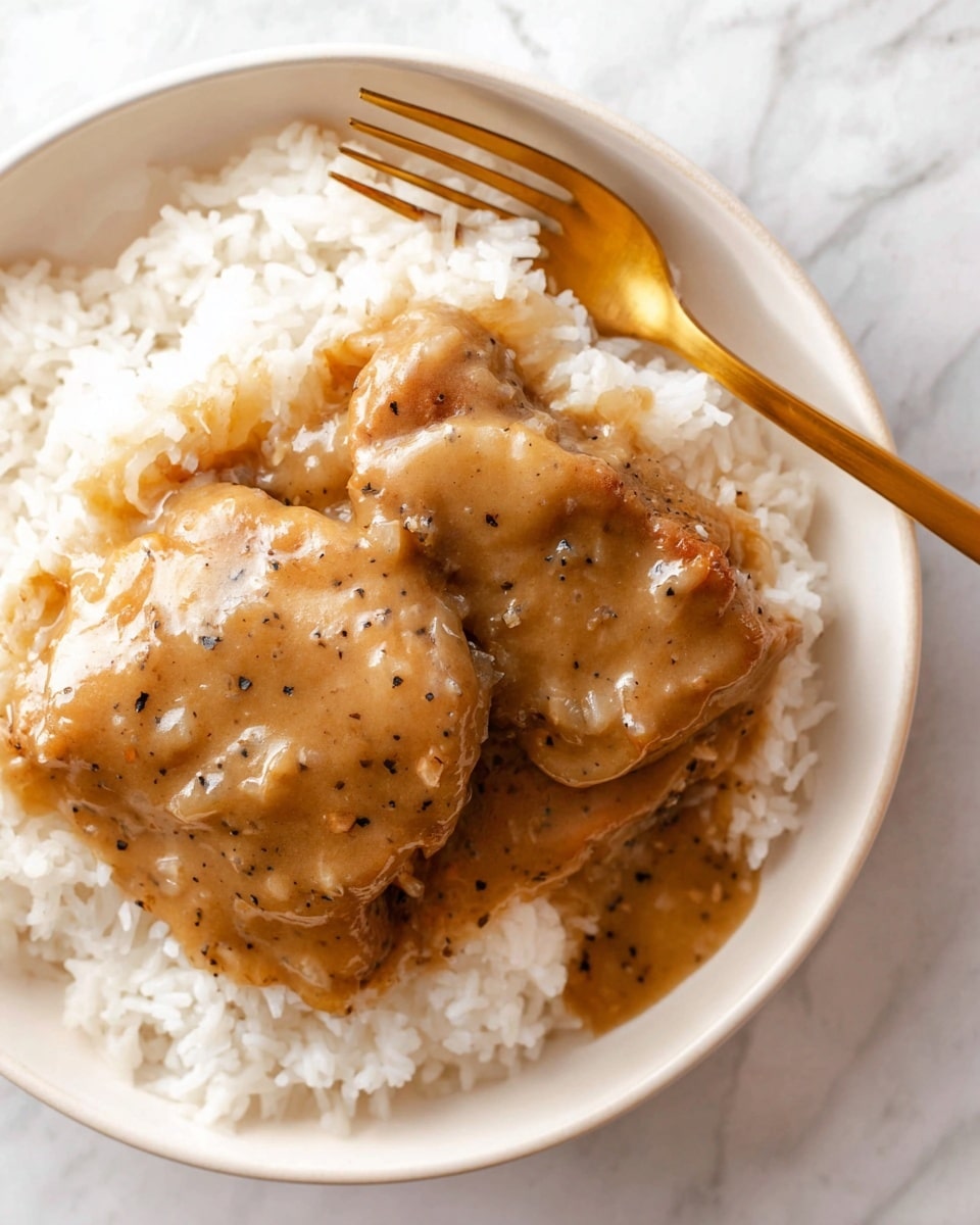 A white bowl holds a bed of white rice at the bottom layer, soft and fluffy. On top, two pieces of brown cooked meat covered in a thick light brown gravy with visible small bits of cooked onion and black pepper specks. The gravy coats the meat and spreads slightly onto the rice. A shiny gold fork rests on the right side of the bowl, touching the rice and sauce. The whole scene is set on a white marbled surface. photo taken with an iphone --ar 4:5 --v 7