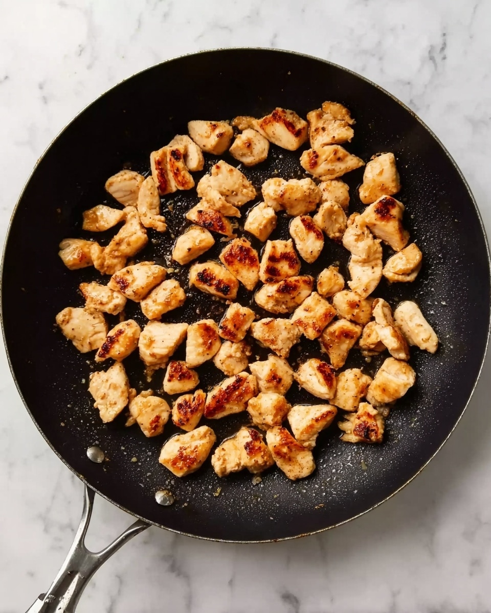 A black frying pan filled with small golden brown pieces of cooked chicken spread evenly inside. The chicken pieces have a slightly crispy texture with some browned spots and are arranged loosely, not overlapping much. The pan sits on a white marbled surface, showing the pan handle pointing towards the lower left side of the image. The lighting highlights the warm colors of the chicken and the pan's black surface photo taken with an iphone --ar 4:5 --v 7