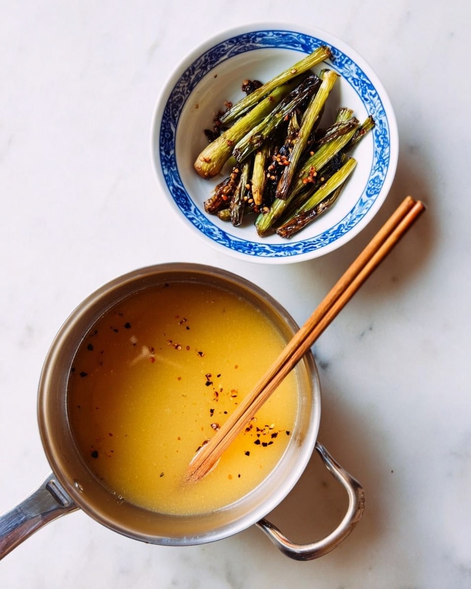 The image shows a small white bowl with blue patterns on the inside rim, filled with grilled green onions that are slightly charred and dark spots of seasoning or spices scattered around them. Below the bowl is a silver metal saucepan containing a golden yellow broth with a smooth texture, and a pair of wooden chopsticks resting inside the saucepan. Both items are placed on a white marbled surface. photo taken with an iphone --ar 4:5 --v 7