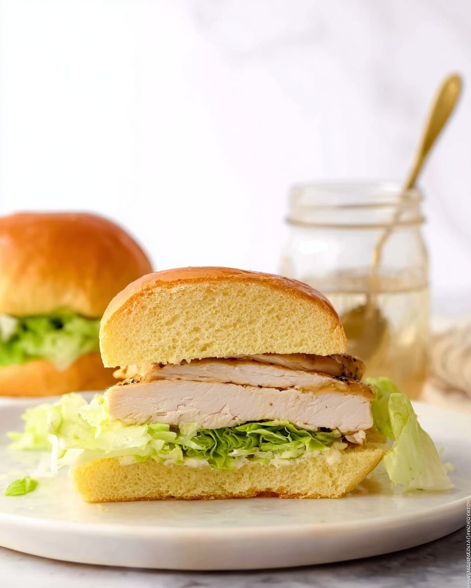 A sandwich is shown in close-up, cut in half and placed on a white plate with a white marbled texture underneath. The sandwich has three main layers: a soft golden-brown bun on the top and bottom, a thick piece of light beige cooked chicken in the middle, and a layer of pinkish-green fresh lettuce between the chicken and the top bun. In the background, there is a clear glass jar with a spoon inside, partially blurred. The sandwich looks fresh and simple, with a focus on the textures of the bun, chicken, and lettuce. Photo taken with an iphone --ar 4:5 --v 7