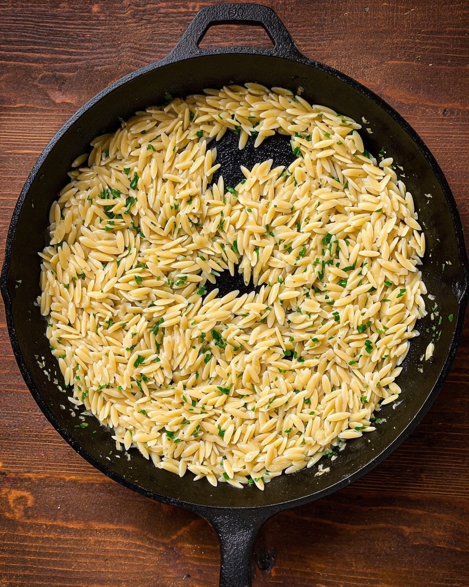 A black cast iron pan filled with a single layer of pale golden orzo pasta cooked and lightly coated with green herb bits, mainly concentrated on top and scattered throughout. The pasta is arranged loosely, showing the dark pan surface in the center and around the edges. The pan handle is visible, and the background is a brown wooden surface. Photo taken with an iphone --ar 4:5 --v 7