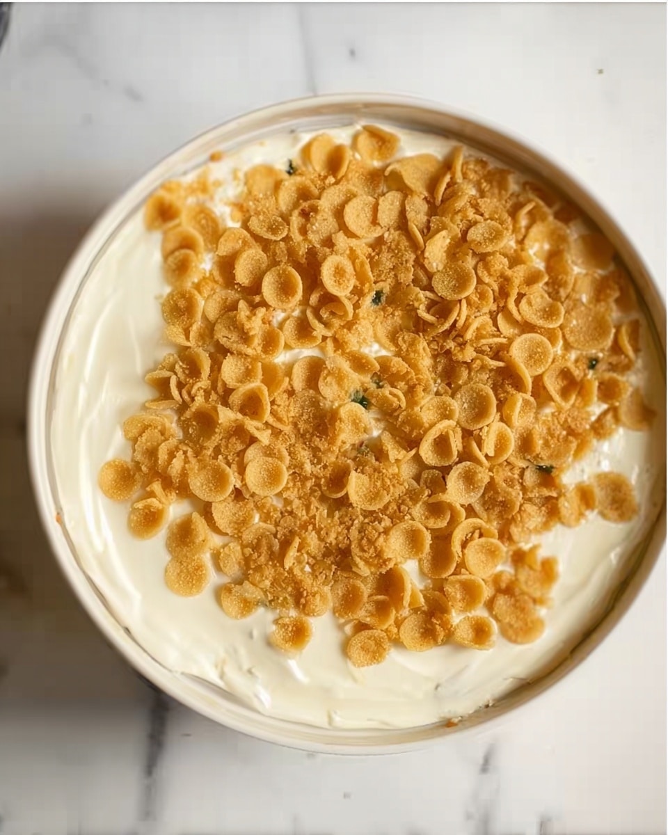 The image shows a close-up view of a dish in a white bowl placed on a white marbled surface. The dish has two main layers visible from the top: a thick, creamy white base layer that looks smooth and soft, and on top of that, there is a layer of small, round, golden-brown pasta or cereal pieces covering the surface evenly. The contrast between the creamy white base and the golden pieces creates a simple but distinct look. Photo taken with an iphone --ar 4:5 --v 7