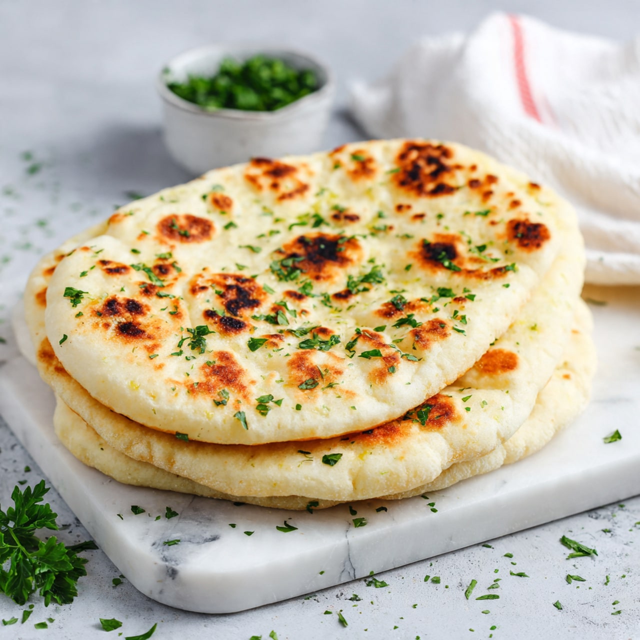 A close-up view of soft, round naan bread with a light golden color and slightly chewy texture, showing small brown charred spots in circular patches on the surface. The naan is stacked in layers, with one torn piece held up revealing the fluffy inside. Small green herb sprinkles decorate the naan, and it is set on a white plate placed against a white marbled background. Photo taken with an iphone --ar 4:5 --v 7