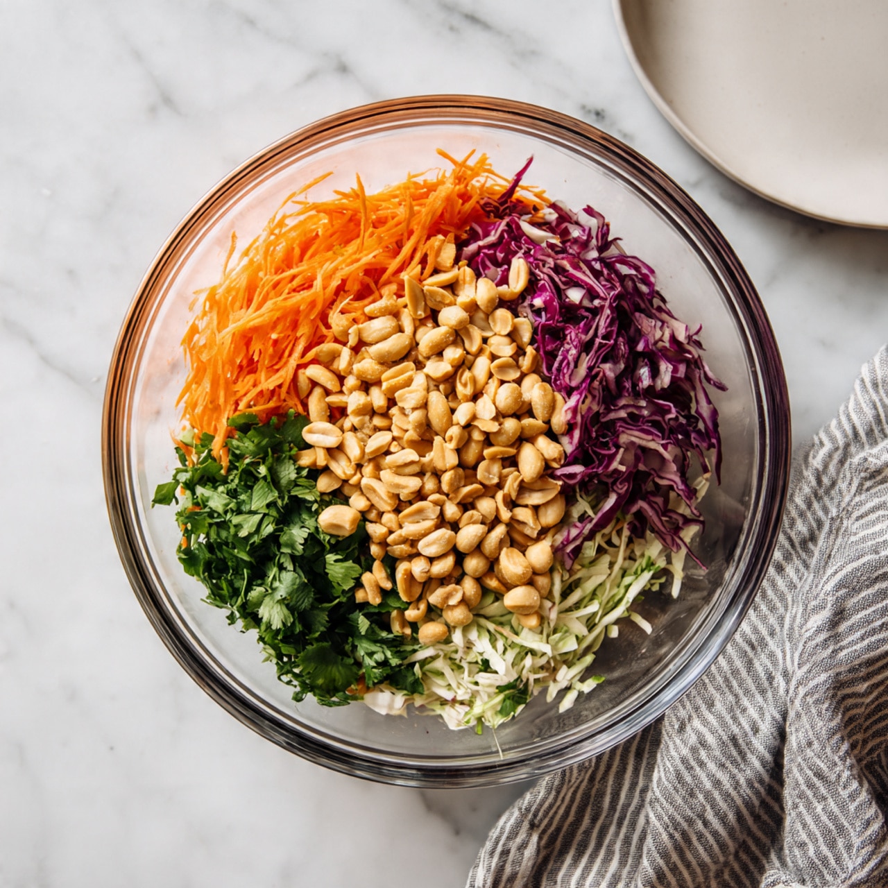 A clear glass bowl sits on a white marbled surface, filled with four distinct layers of colorful ingredients arranged side by side. Starting from the top right, there is a bright orange layer of shredded carrots with a thin, soft texture. Below this, on the bottom right, is a mix of thinly shredded white and purple cabbage, showing a crisp and slightly curly texture. To the left of the cabbage, at the bottom left of the bowl, is a pile of finely chopped fresh green herbs with a leafy, moist appearance. The center of the bowl is topped with a generous handful of light tan peanuts, adding a smooth, rounded contrast to the other layers. A clean white plate is partially visible in the top right corner, and a gray and white striped cloth lays on the bottom right corner of the white marbled surface. photo taken with an iphone --ar 4:5 --v 7