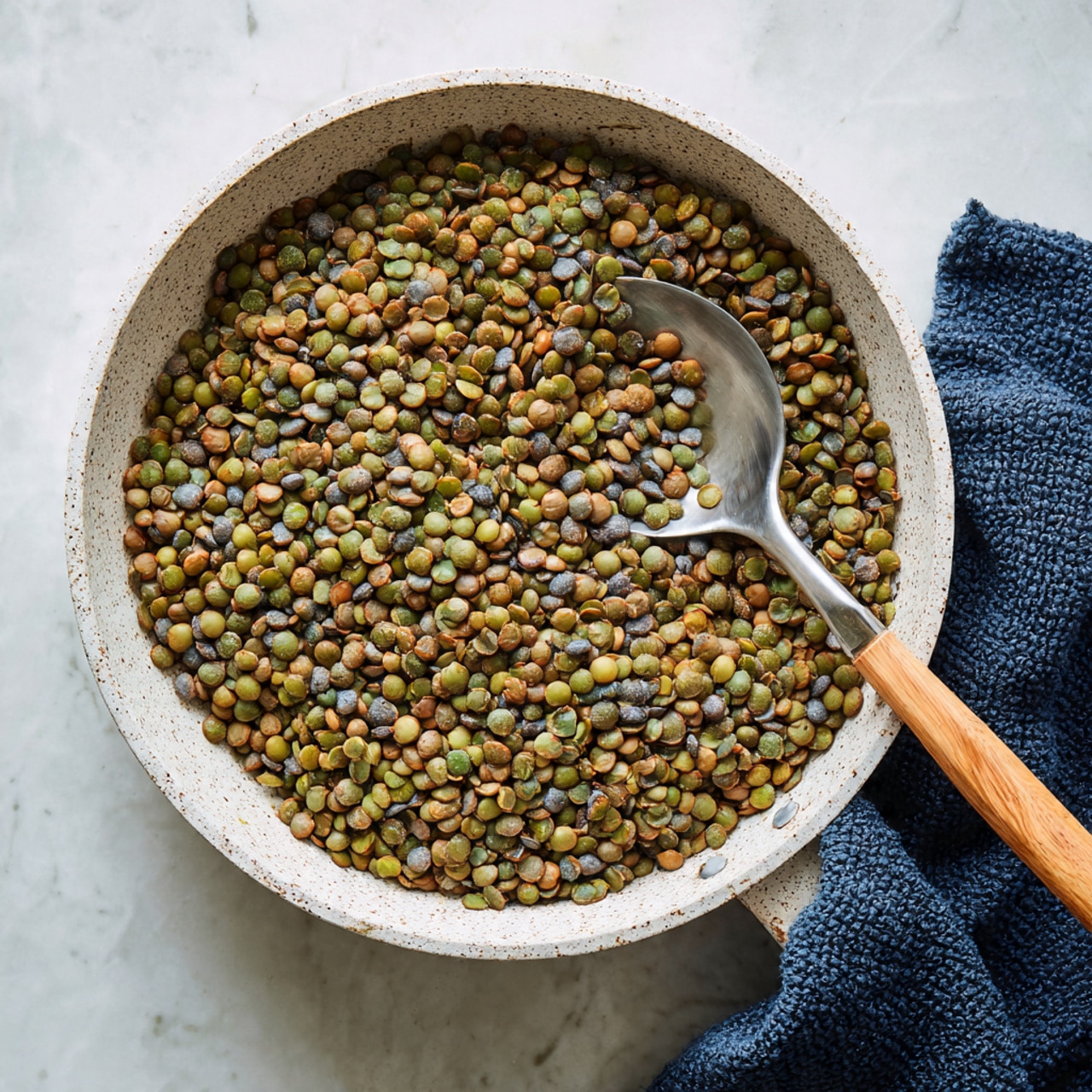 A round white speckled pan filled with cooked green lentils, showing different shades of green and brown with a soft texture. A shiny metal spoon with a light wooden handle lies inside the pan, partially covered by the lentils. The pan’s handle is light wood, extending slightly out of frame on a white marbled surface. To the right side, a folded dark blue knitted cloth rests next to the pan. The photo taken with an iphone --ar 4:5 --v 7