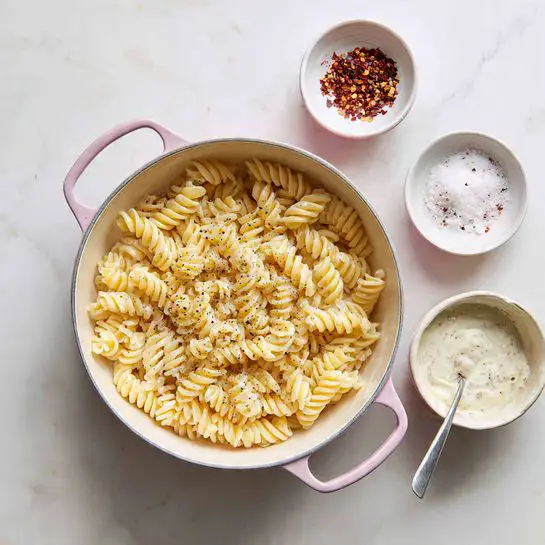The image shows a pot filled with creamy macaroni and cheese, with a wooden spoon lifting a portion of the pasta in the center. The macaroni noodles are thick and curly, coated in a smooth, light yellow cheese sauce with a few specks of black pepper on top. The pot has orange handles and sits on a white marbled surface. In the background, a yellow cloth is partially visible, adding a warm tone to the scene. The focus is on the spoonful of macaroni and cheese, highlighting the creamy texture and the dense layer of noodles. Photo taken with an iphone --ar 4:5 --v 7