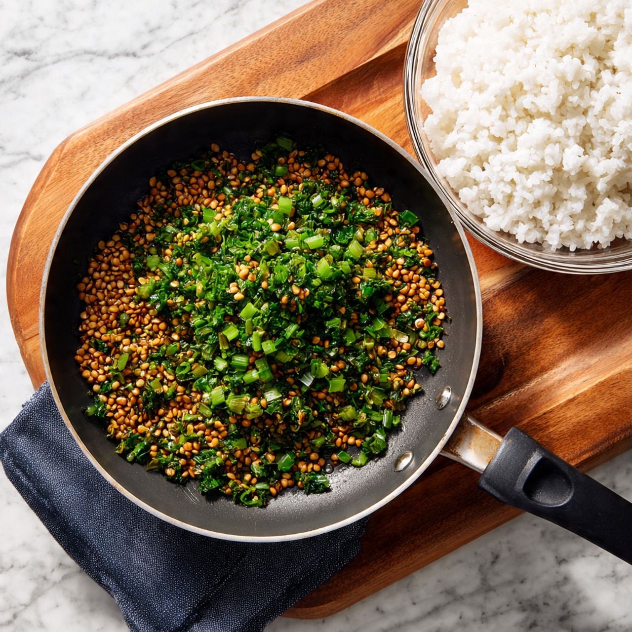 A black pan with a silver rim and a black handle sits on a folded dark blue cloth on top of a round wooden board, all placed on a white marbled surface. Inside the pan are small golden brown seeds scattered around the bottom along with chopped green pieces evenly spread mainly in the center. To the upper right of the pan, a clear glass bowl shows white cooked rice grains. Photo taken with an iphone --ar 4:5 --v 7