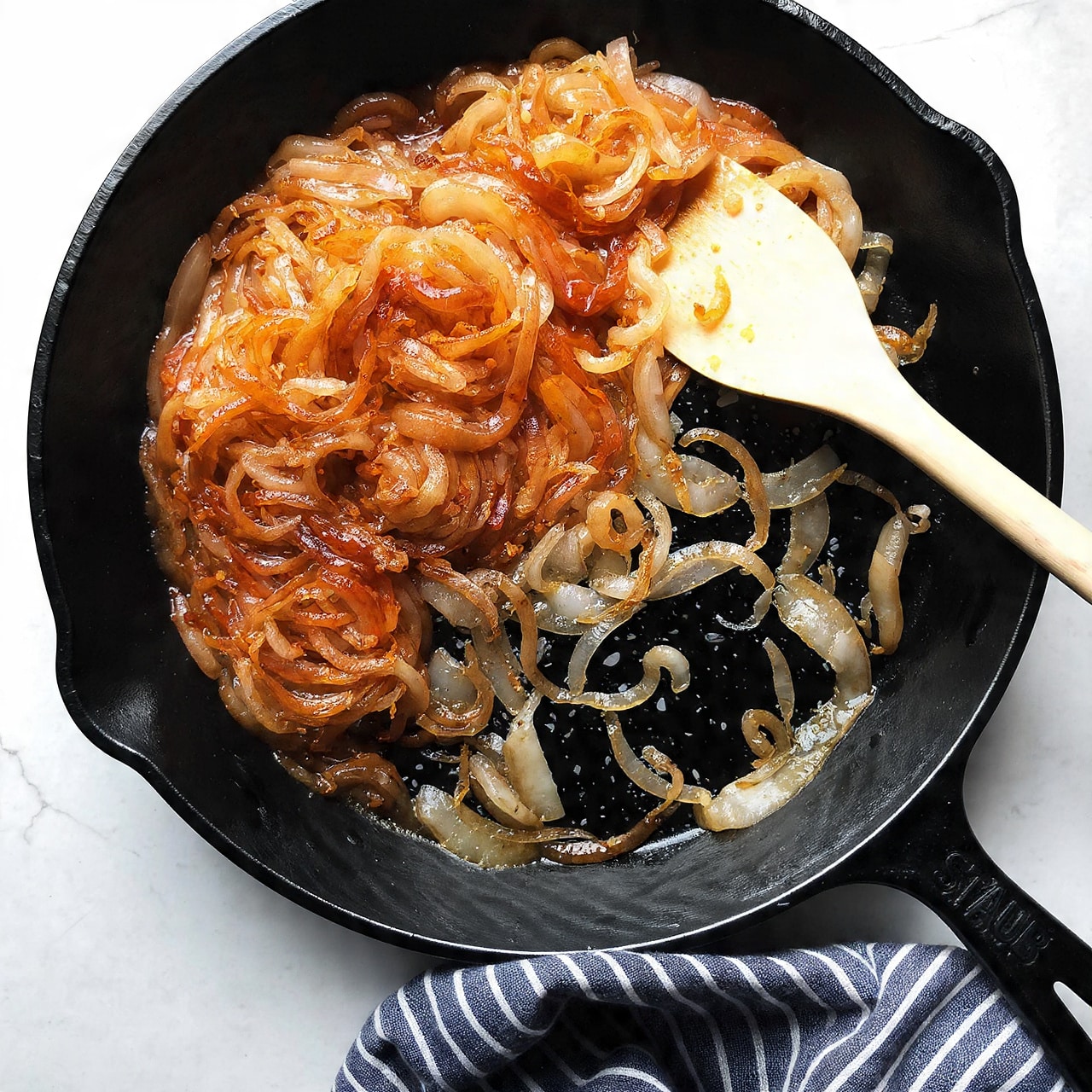 A black cast iron pan holds a single layer of caramelized onions, showing shades of golden brown and light amber with some darker browned parts. The onions are soft and slightly translucent with thin curled strips unevenly spread. A light wooden spatula rests on top of the onions near the right side of the pan. The pan sits on a white marbled surface with a crumpled dark gray and white striped cloth near its lower edge. The handle of the pan is dark blue with the word