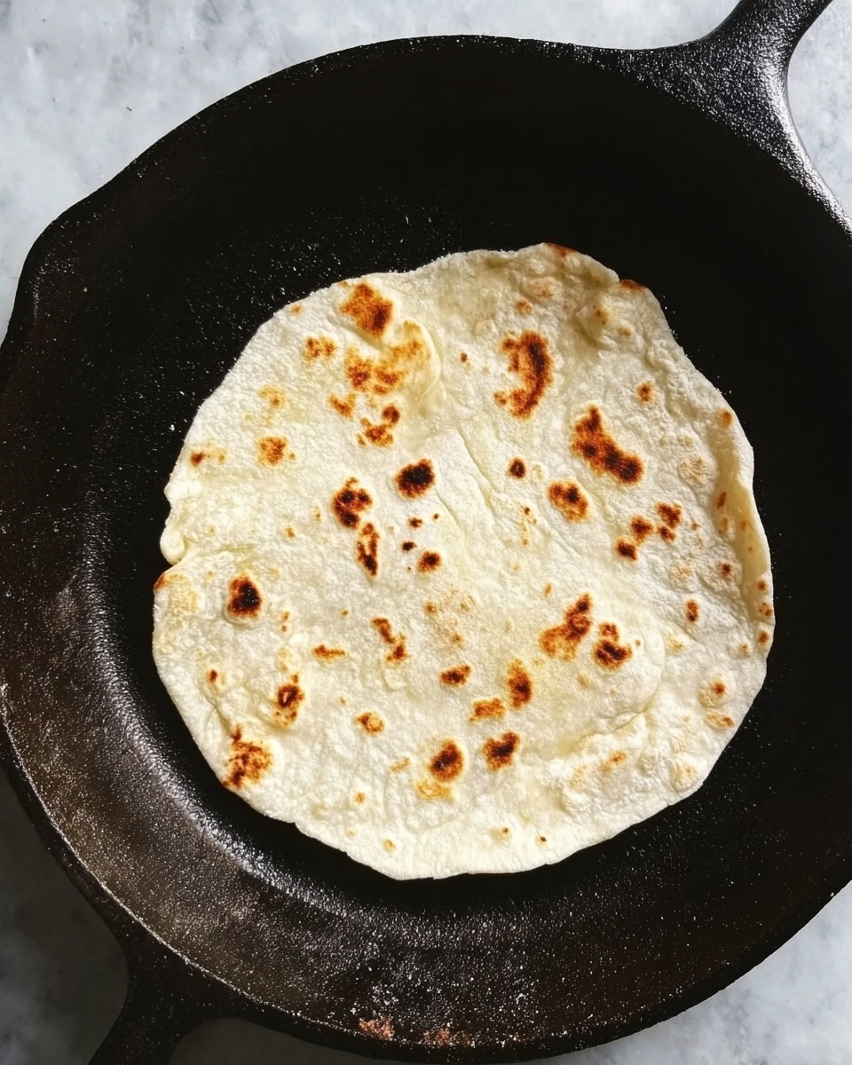 A round, thin flatbread with small brown spots where it was cooked lies flat inside a black cast iron pan. The flatbread is pale white with uneven edges and some slight puffiness. The pan has a rough, dark surface with slight seasoning marks visible around the bread. The background is a white marbled texture. photo taken with an iphone --ar 4:5 --v 7