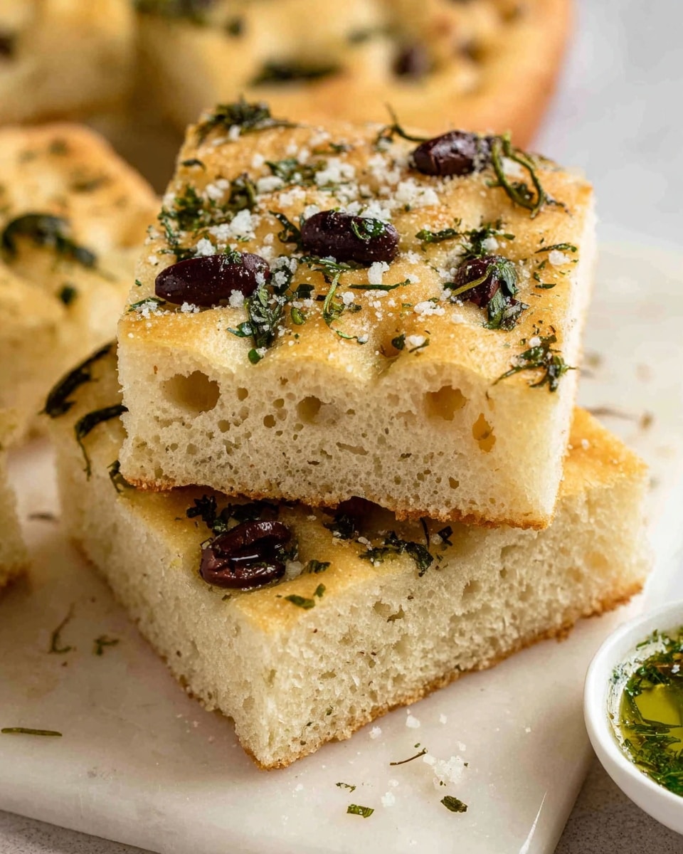 The image shows two thick square pieces of focaccia bread stacked on top of each other on a white marbled surface. Each piece has a light golden-brown top with a soft, airy, and slightly spongy texture inside. The top layer is sprinkled with chopped dark olives, coarse salt crystals, and green herbs, giving it a fresh and slightly rough look. Around the main stack, more pieces of focaccia can be seen, with similar toppings and texture. A small white bowl with a green sauce or dip is partially visible to the right, adding a hint of color contrast. Photo taken with an iphone --ar 4:5 --v 7