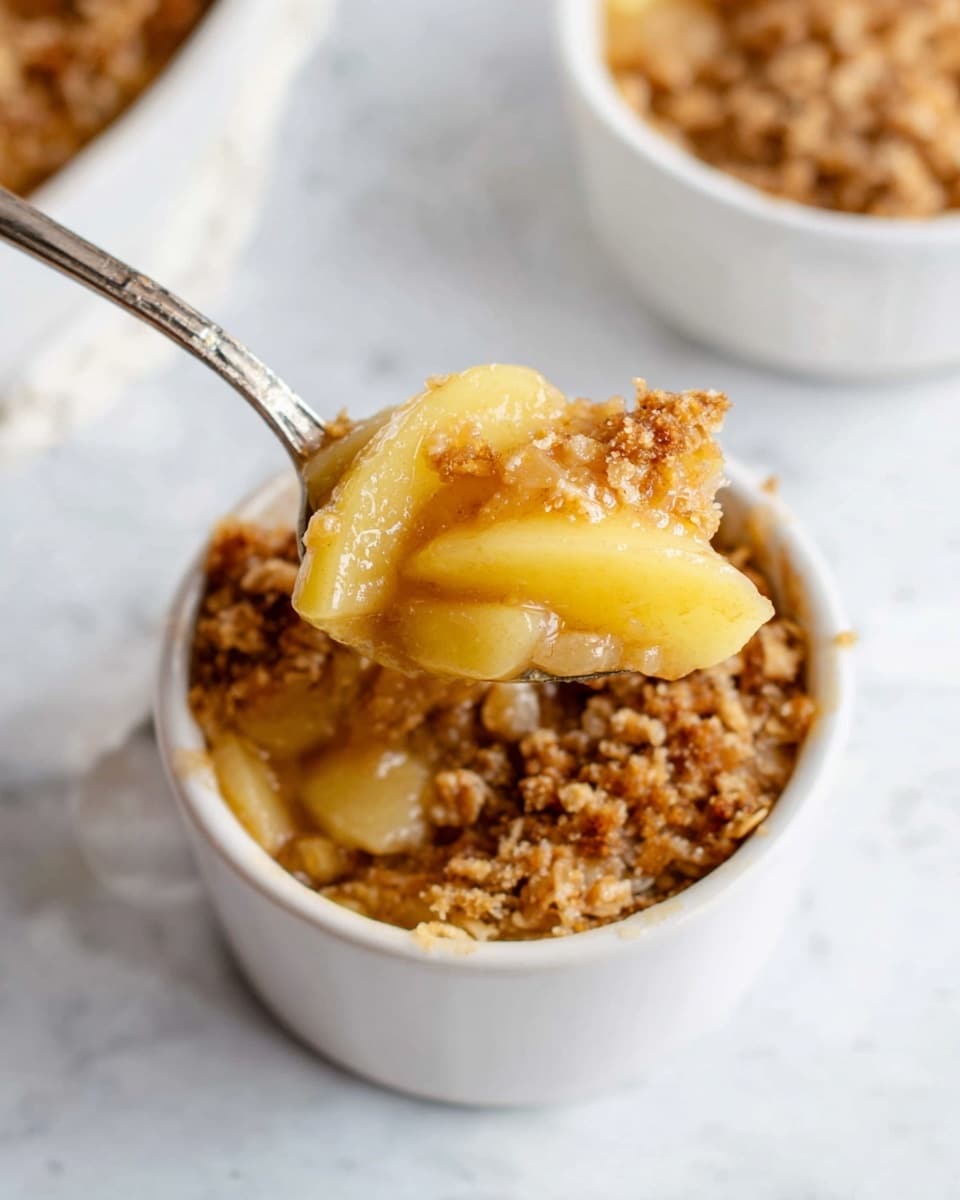 The image shows a small white bowl filled with a dessert that has two main layers: a bottom layer of soft yellow apple slices with a slightly glossy, cooked texture and a top layer of golden brown crumbly oat mixture with a grainy texture. A silver spoon lifts a portion of the dessert, showing the apple slices on the bottom and the oat topping on top. The bowl is placed on a white marbled surface with another similar white bowl partially visible nearby. Photo taken with an iphone --ar 4:5 --v 7