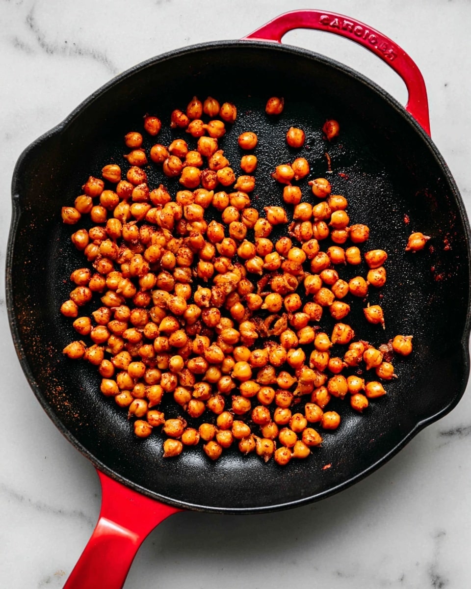 A black cast iron pan with a bright red handle and side grip is filled with a single layer of roasted chickpeas. The chickpeas are golden brown with a slightly shiny, crispy texture, spread mostly across the center and left side of the pan, leaving the right side mostly empty. The pan sits on a white marbled surface that provides a clean, bright contrast to the dark pan and colorful chickpeas. photo taken with an iphone --ar 4:5 --v 7