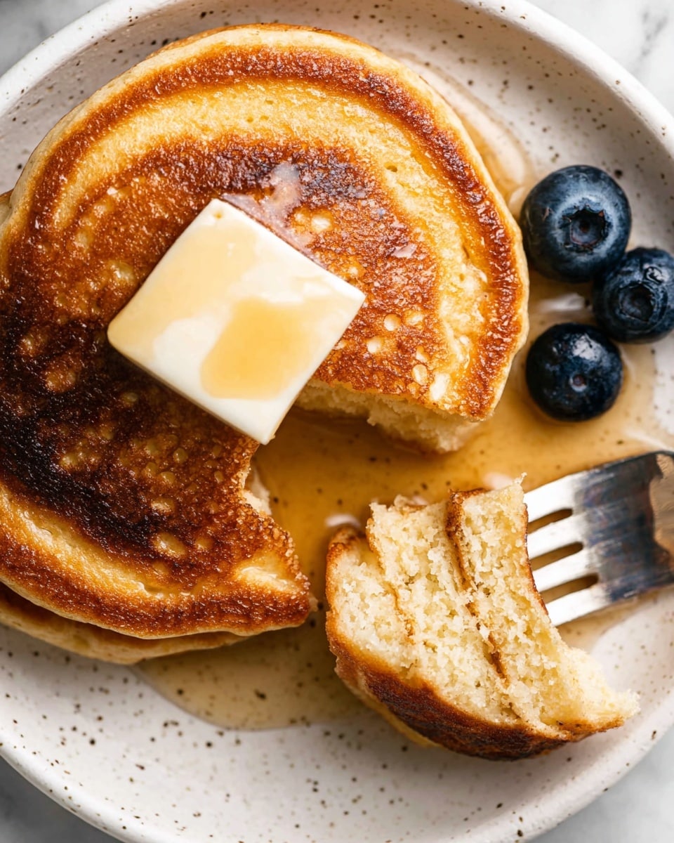 The image shows a close-up of two golden brown pancakes on a white plate with small dark speckles. The pancake in the center has a shiny square pat of melting butter on top with honey or syrup glistening around it. A section of the same pancake is broken off and held by a silver fork, showing the fluffy, light beige inside texture. Near the pancakes, a few fresh blueberries add a dark blue contrast. The whole scene is set on a white marbled surface. Photo taken with an iphone --ar 4:5 --v 7