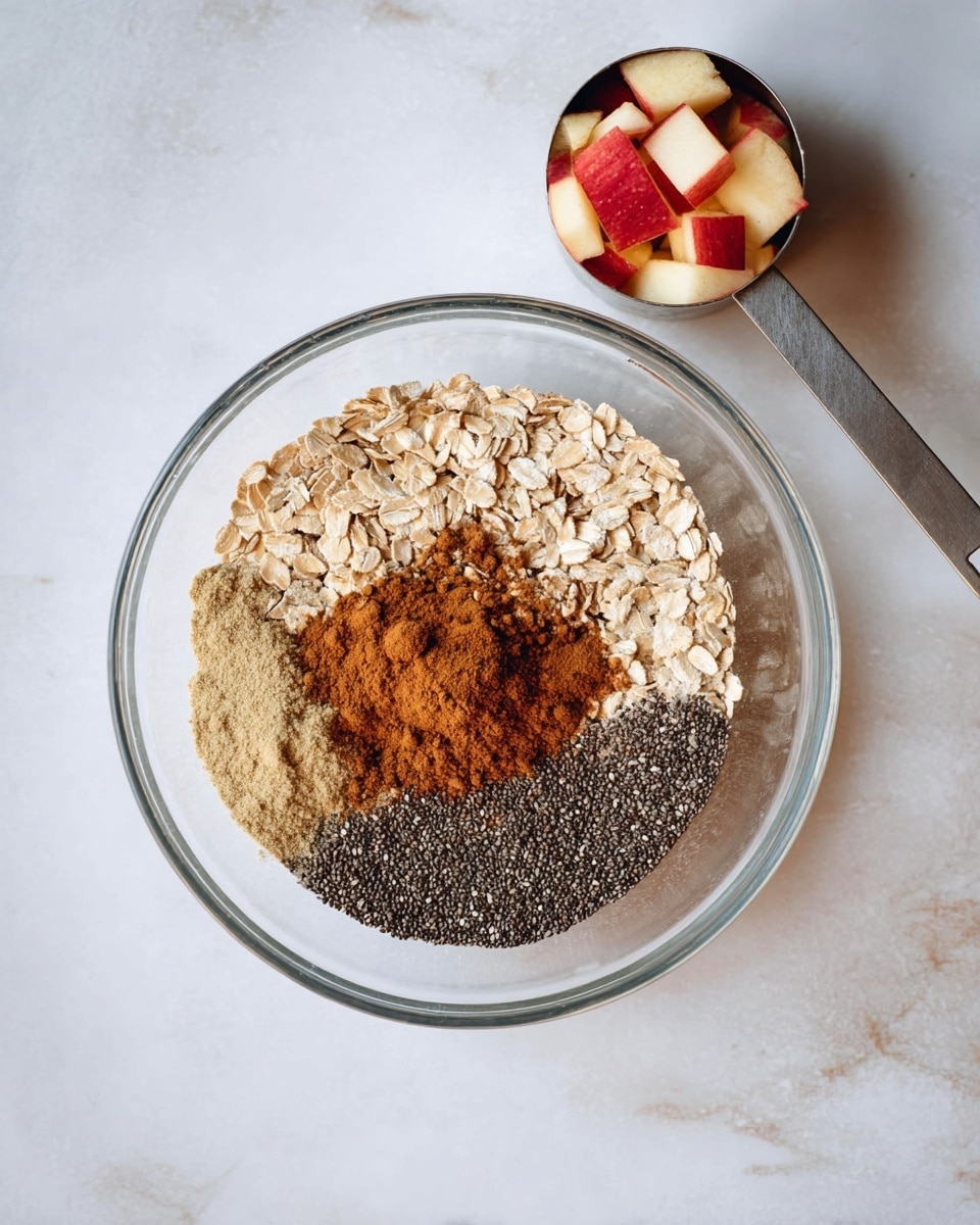 A clear glass bowl holds four distinct layers of dry ingredients placed side by side in a circular arrangement: light beige oats on the top left, a heap of brown cinnamon powder in the center, a small mound of pale beige ginger powder on the lower left next to the cinnamon, and tiny black chia seeds covering the right half of the bowl. Above this bowl, there is a metallic measuring cup filled with red and white chopped apple pieces. The background is a white marbled surface. Photo taken with an iphone --ar 4:5 --v 7