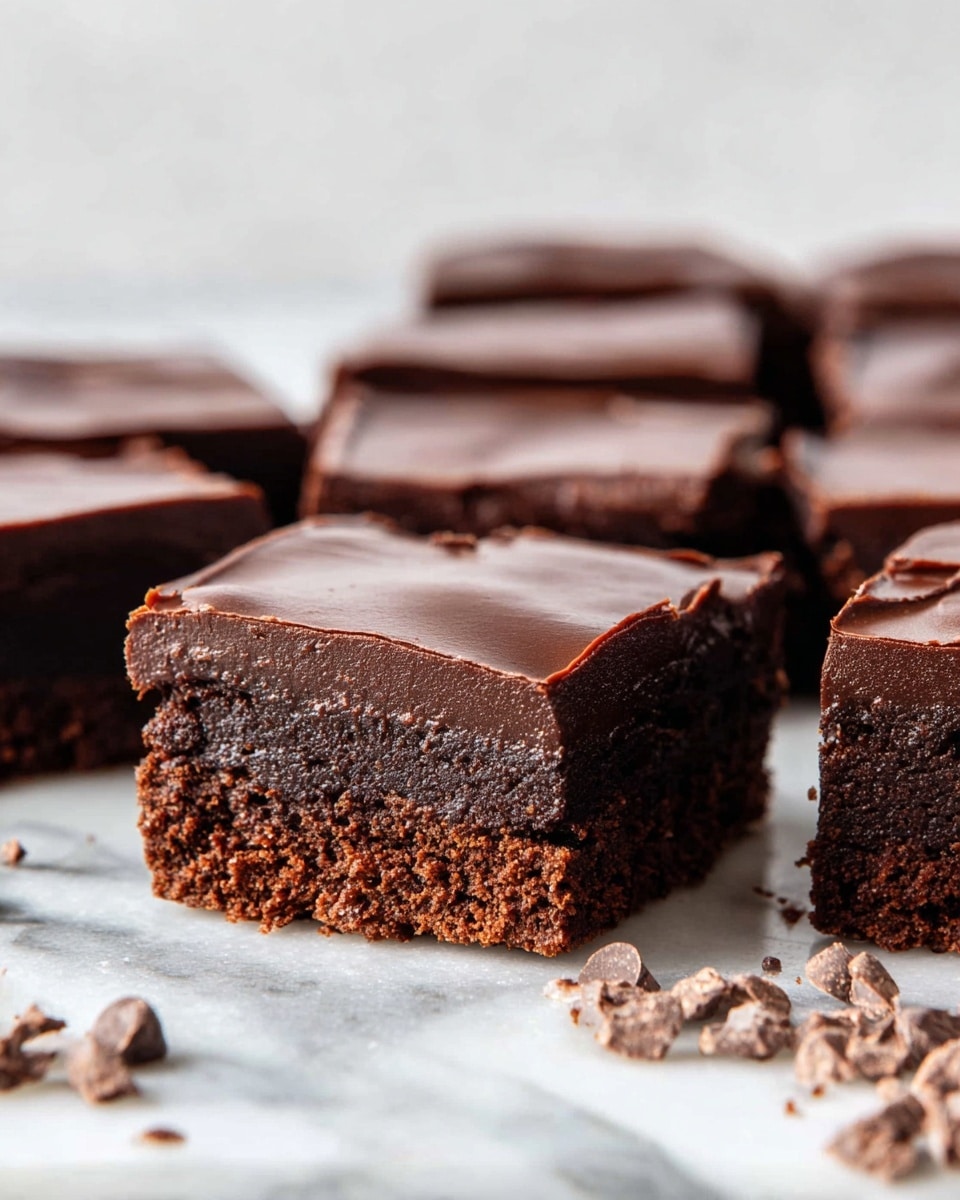 The image shows a close-up of several square chocolate brownies arranged neatly on a white marbled surface. Each brownie has two layers: a dense, slightly rough, dark brown bottom layer that looks soft and fudgy, and a smooth, shiny, darker brown top layer of chocolate frosting that evenly covers each piece. In front of the brownies, there are small scattered pieces of chocolate or cocoa nibs adding texture to the scene. The background is a simple white marbled texture with soft lighting that highlights the moist texture of the brownies. Photo taken with an iphone --ar 4:5 --v 7