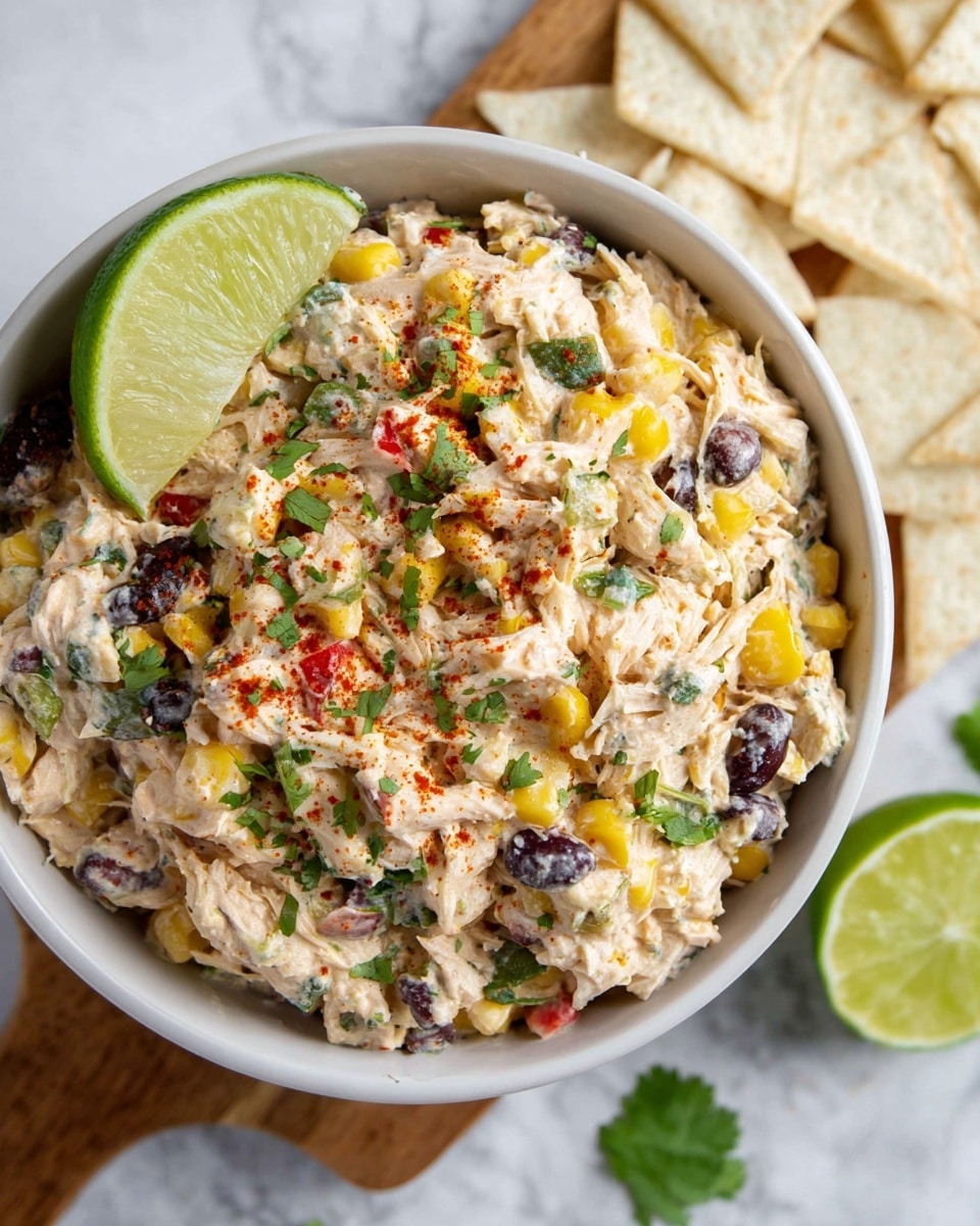 The image shows a white bowl filled with a mixed salad that has multiple layers and colors, placed on a white marbled surface. The top layer has shredded cream-colored chicken pieces mixed with yellow corn kernels and black beans, all coated with a creamy dressing. There are small pieces of red and green vegetables, likely bell peppers and herbs, scattered throughout. The dish is garnished with chopped fresh green cilantro and light red paprika powder sprinkled on top. On the side of the bowl, a bright green lime wedge rests against the salad. Next to the bowl, white triangular crackers or chips are neatly arranged on a wooden board partly visible at the top. The photo is clear and bright, taken with an iphone --ar 4:5 --v 7