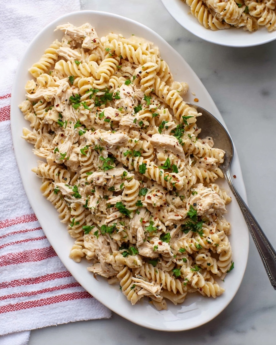 A white oval plate filled with creamy rotini pasta mixed with shredded light beige chicken, all coated in a soft cream sauce flecked with black pepper and tiny red spices, sprinkled with fresh bright green parsley leaves for garnish. The pasta shows ridged spiral shapes with a slightly glossy texture, and the chicken is shredded into thin strips blending evenly through the pasta. A large silver serving spoon rests on the top right edge of the plate. The white marbled background has a subtle pattern, with a white cloth with red stripes at the bottom left corner and a partial view of another white bowl of the same pasta above on the right side. photo taken with an iphone --ar 4:5 --v 7