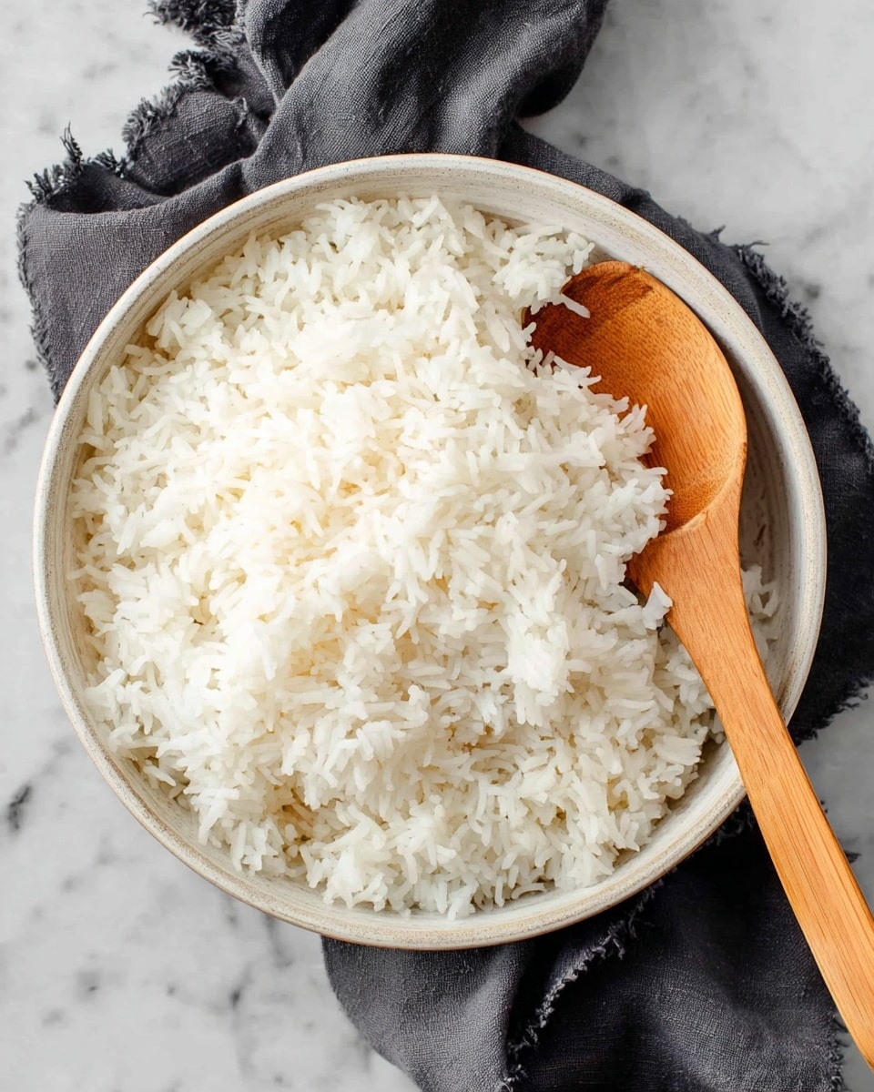 A bowl filled with fluffy white cooked rice, each grain separated and soft in texture, filling the bowl almost to the top. A wooden spoon is placed inside the rice on the right side, its smooth light brown color contrasting with the white rice. The bowl is white with a subtle texture, sitting on a white marbled surface. A dark grey cloth napkin with a slight crease lies beside the bowl, adding a soft fabric detail to the scene. Photo taken with an iphone --ar 4:5 --v 7