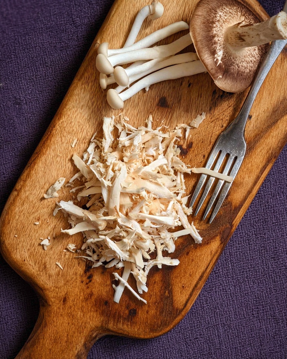 The image shows a wooden cutting board with a pile of shredded light beige mushrooms in the center. On the top side of the board, there is a small bunch of whole white mushrooms with thin stems and a single large mushroom cap with a brown and white color. A silver fork rests on the bottom right edge of the board, with some mushroom pieces caught between its prongs. The cutting board is placed on a purple textured fabric, and the top view highlights the natural wood grains and the varied textures of the mushrooms. photo taken with an iphone --ar 4:5 --v 7
