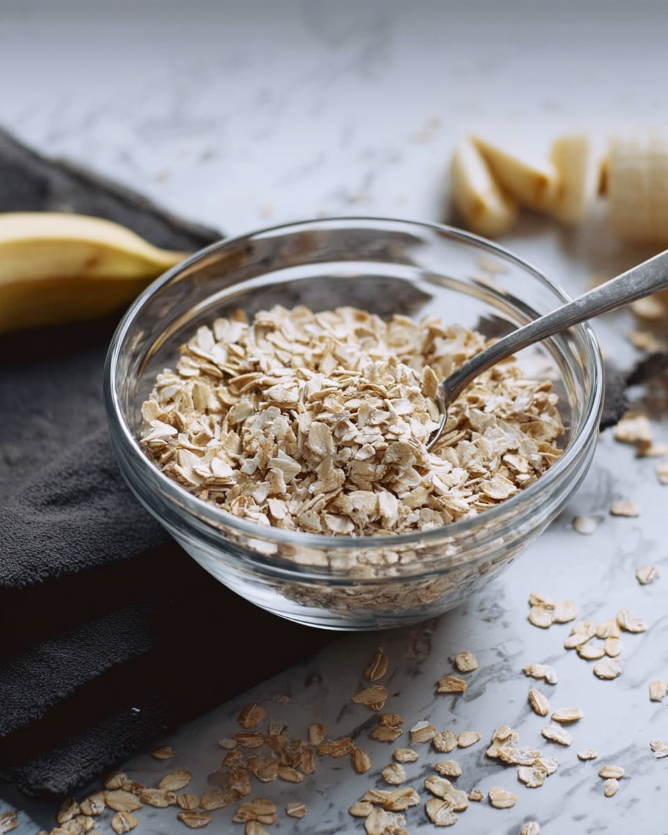 The image shows a clear glass bowl filled with dry, beige rolled oats. A metal spoon is placed inside the bowl, with its handle sticking out, resting on the oats. Behind the bowl, there is a peeled banana sliced into pieces, slightly out of focus. The bowl sits on a dark cloth, and the surface below is a white marbled texture. Some scattered oats are visible around the bowl, adding a casual, natural feel. Photo taken with an iphone --ar 4:5 --v 7