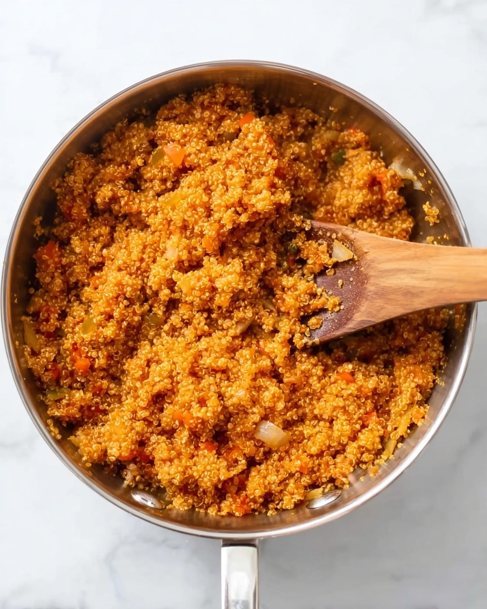 A metal pan filled with cooked quinoa that has a deep orange color, mixed evenly with small pieces of soft vegetables such as onions and peppers. The quinoa has a fluffy and slightly moist texture, with tiny spiral-shaped grains visible throughout. A wooden spoon is placed inside the pan on the right side, partially covered in the quinoa mixture. The pan is sitting on a white marbled surface. photo taken with an iphone --ar 4:5 --v 7