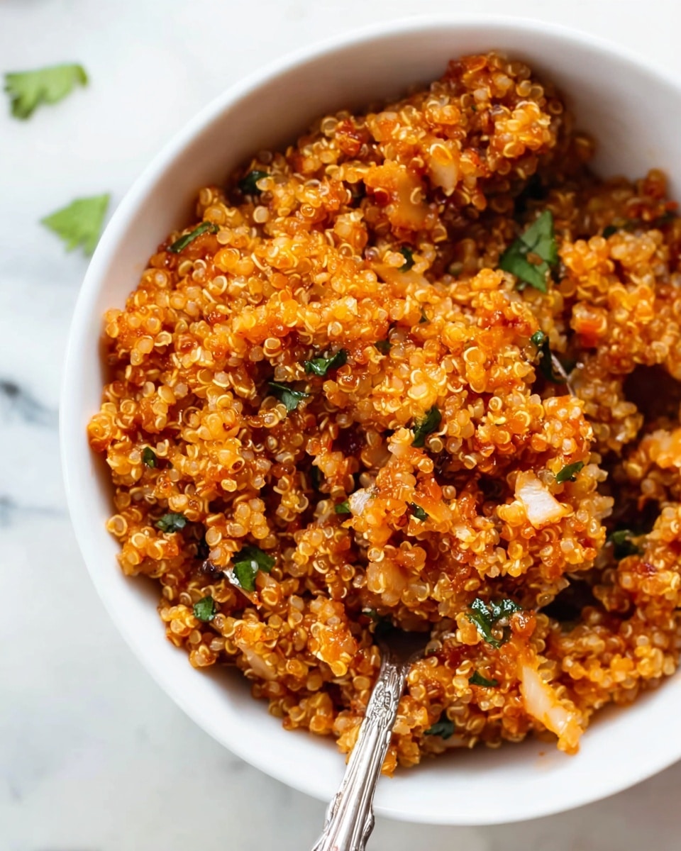 A close-up view of a white bowl filled with three layers of cooked quinoa mixed with small pieces of onion and green herbs, all coated in a reddish-orange sauce giving a warm tone to the dish. The quinoa grains are fluffy and separated with visible small curls, and the small vegetable pieces add texture and color contrast. A vintage silver spoon rests inside the bowl, partly covered by quinoa. The bowl sits on a white marbled surface with a few blurred green herb pieces scattered nearby. Photo taken with an iphone --ar 4:5 --v 7