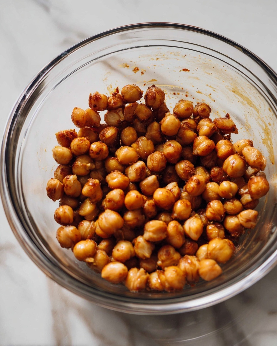 The image shows a clear glass bowl filled with one layer of golden brown roasted chickpeas that have a slightly shiny, textured surface, some with darker toasted spots, sitting on a white marbled surface. photo taken with an iphone --ar 4:5 --v 7
