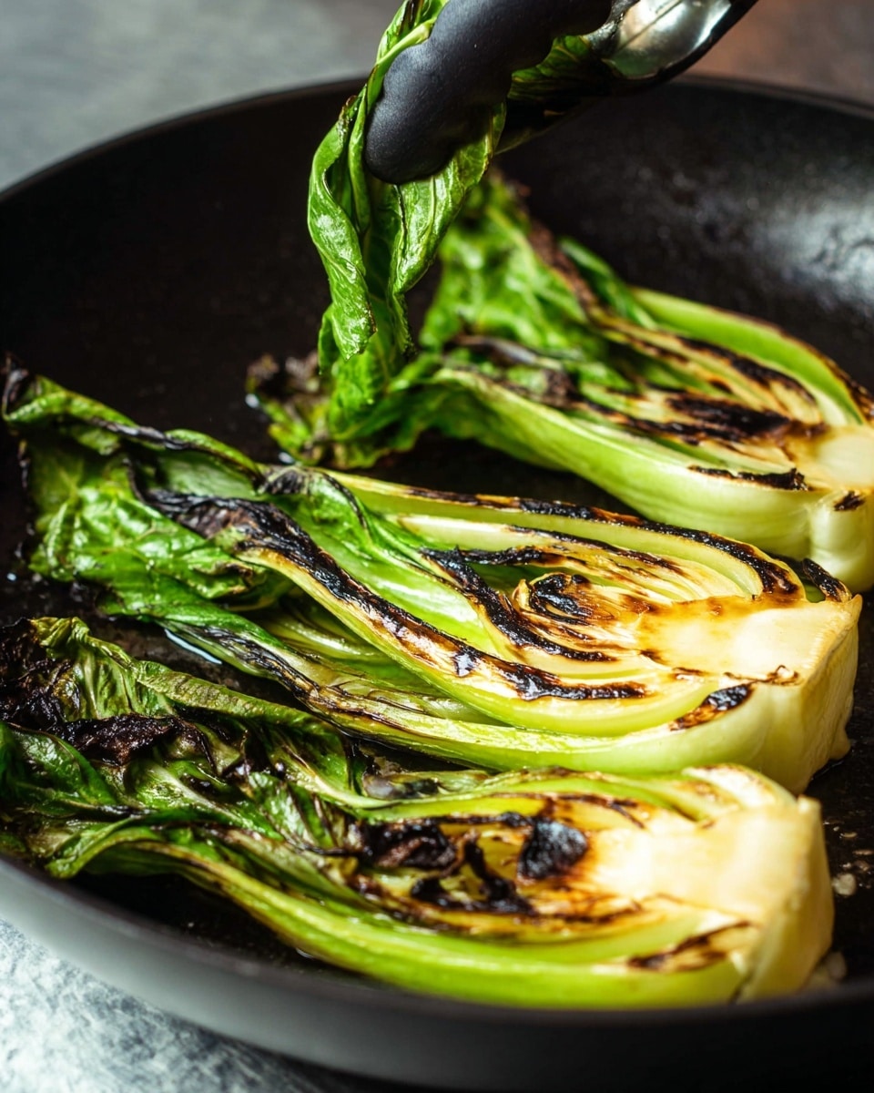 The image shows three pieces of grilled bok choy in a black pan. Each bok choy is cut in half lengthwise, showing layers of light green and white at the base with darker green leaves on top. The grilled parts have dark brown, almost black, char marks on the edges and parts of the leaves. A woman's hand holding black tongs is lifting one piece slightly. The background beneath the pan is a white marbled texture. photo taken with an iphone --ar 4:5 --v 7