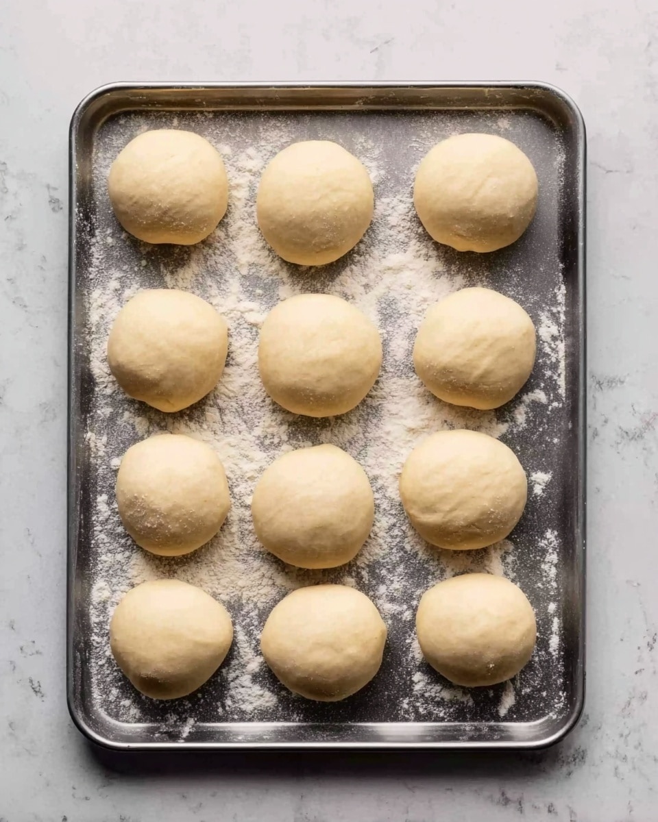 The image shows 12 round dough balls arranged in three rows on a metal baking tray dusted with flour. Each dough ball is smooth with a pale beige color and slightly shiny texture, evenly spaced on the tray. The tray is placed on a white marbled surface, creating a clean and bright background. Photo taken with an iphone --ar 4:5 --v 7