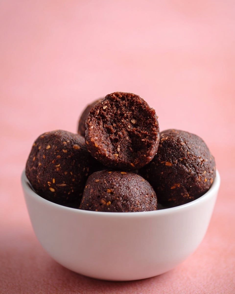 A white bowl filled with six round dark brown balls that look like chocolate bites, with small lighter brown bits inside. One ball is on top, broken in half to show a dense, crumbly inside texture. The background is soft pink and smooth. photo taken with an iphone --ar 4:5 --v 7