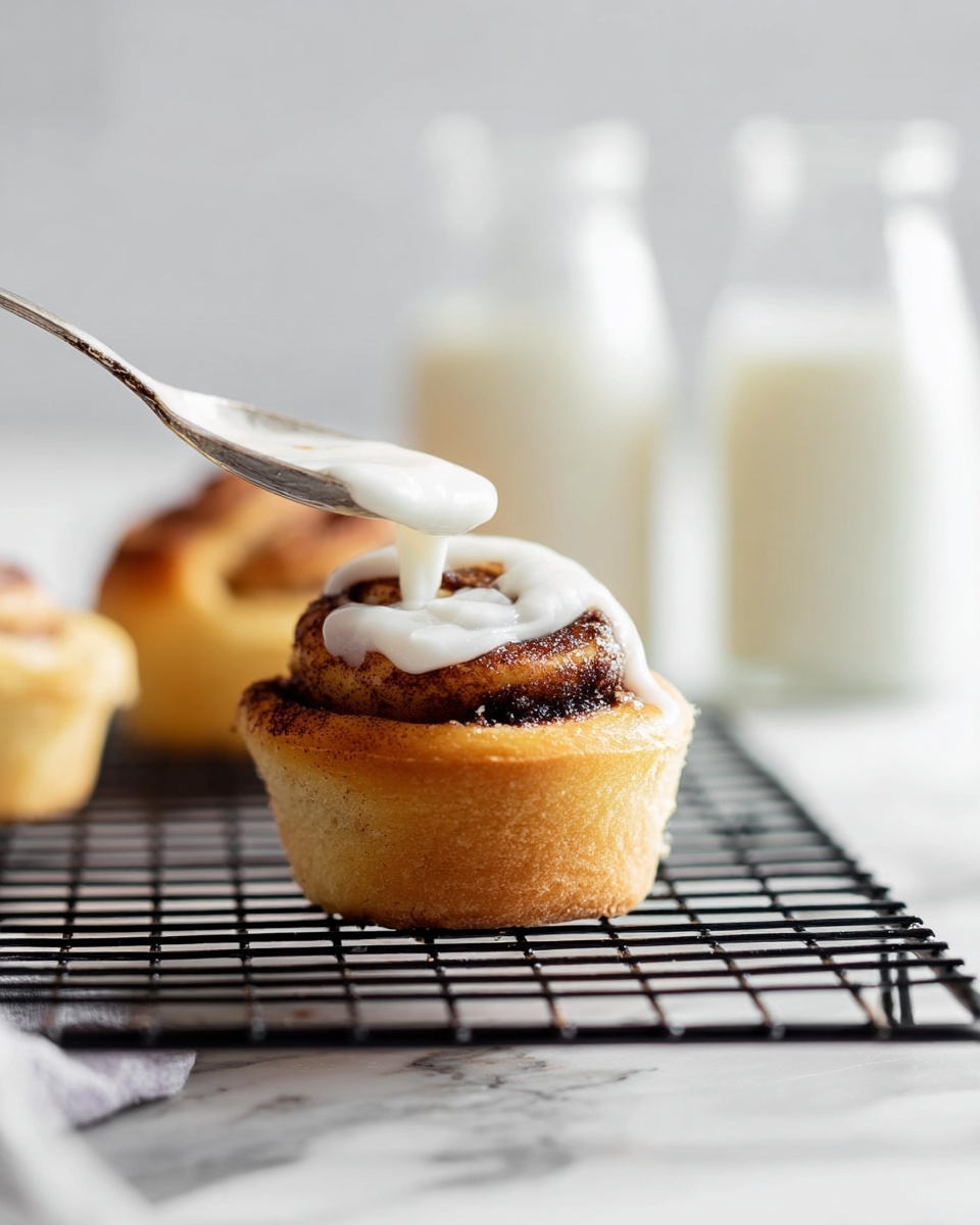 A close-up of a small cinnamon roll sitting on a black wire cooling rack over a white marbled surface, showing one thick base layer of golden-brown dough, a second layer spiraled with cinnamon filling that is dark brown and slightly shiny, and a thick layer of white icing being spread on top with a spoon held by a woman's hand from the left side of the frame. In the blurred background, there are two glass bottles filled with milk, all softly lit with natural lighting. Photo taken with an iphone --ar 4:5 --v 7