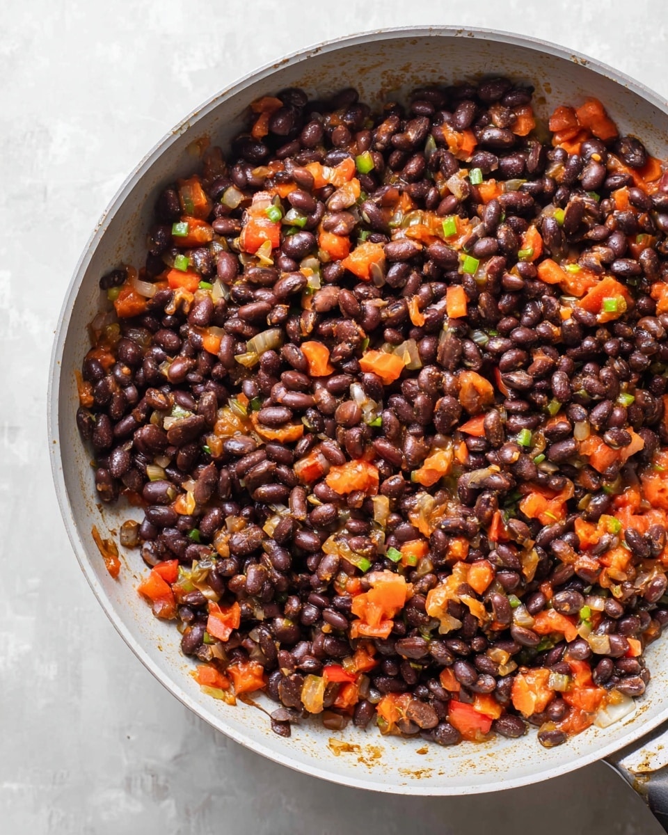 A white pan filled with cooked black beans mixed with small, soft pieces of orange and red tomatoes, translucent bits of onion, and tiny green pepper pieces, all lightly coated in a bit of sauce. The beans are dark purple-brown and evenly spread throughout the pan, creating a textured and colorful mix. The white marbled surface underneath the pan adds a clean, smooth contrast to the dish. photo taken with an iphone --ar 4:5 --v 7